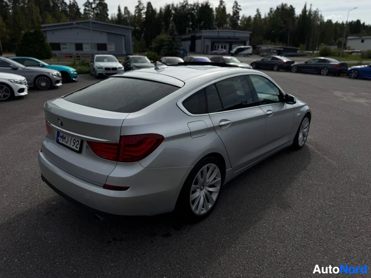 Takaviisto: Hopeanvärinen henkilöauto parkkipaikalla, takaosa ja vasen kylki näkyvissä. Auton takarekisterikilvessä lukee. Taustalla on rakennuksia ja puita
