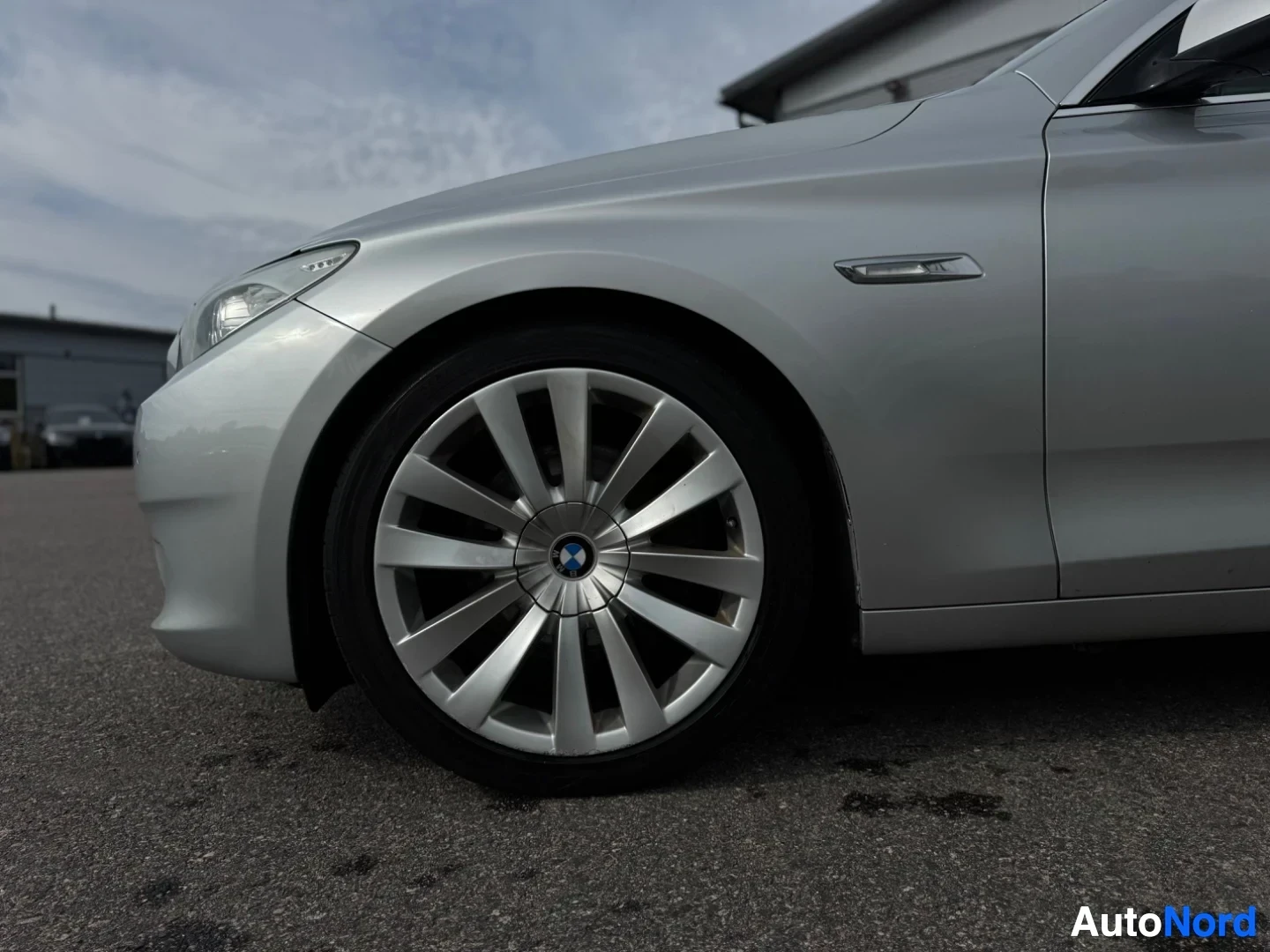 Etuviisto, nokka vasemmalle: Kuva auton etuosan vasemmasta kyljestä ja etupyörästä. Ajovalon yläosa näkyy, samoin etulokasuoja ja sivuvalaisin. Vanteessa on BMW-logo.