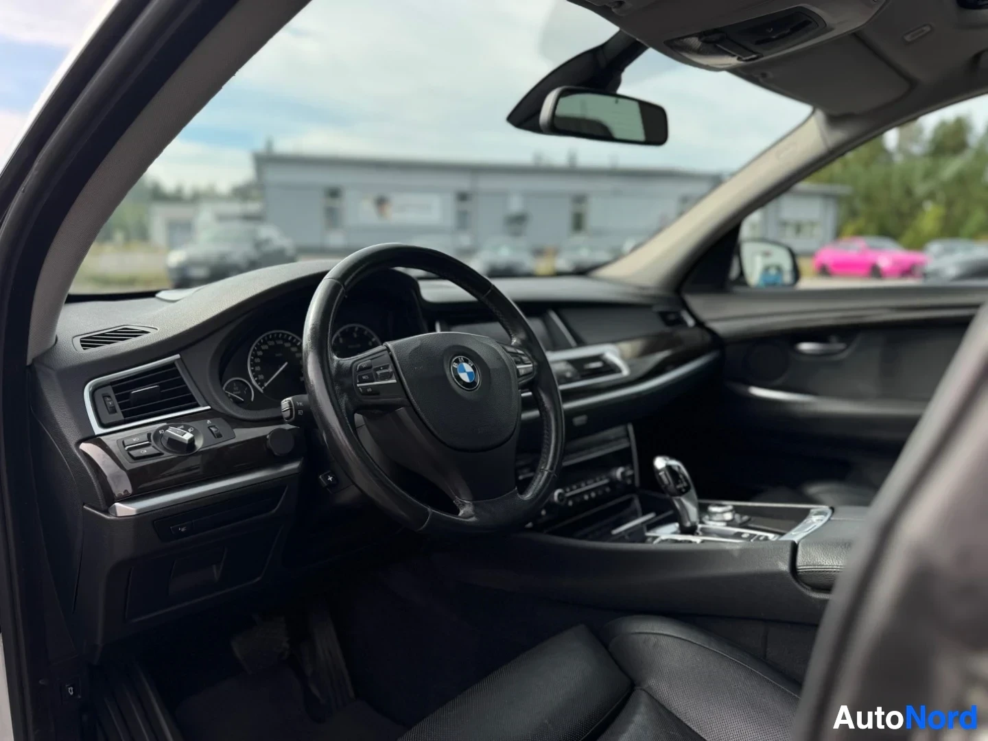 Ohjaamo: Kuvassa näkyy auton kuljettajan puolen ohjaamo, mukaan lukien monitoimiohjauspyörä BMW-logolla, mittaristo ja keskikonsoli. Taustalla näkyy rakennuksia ja puita