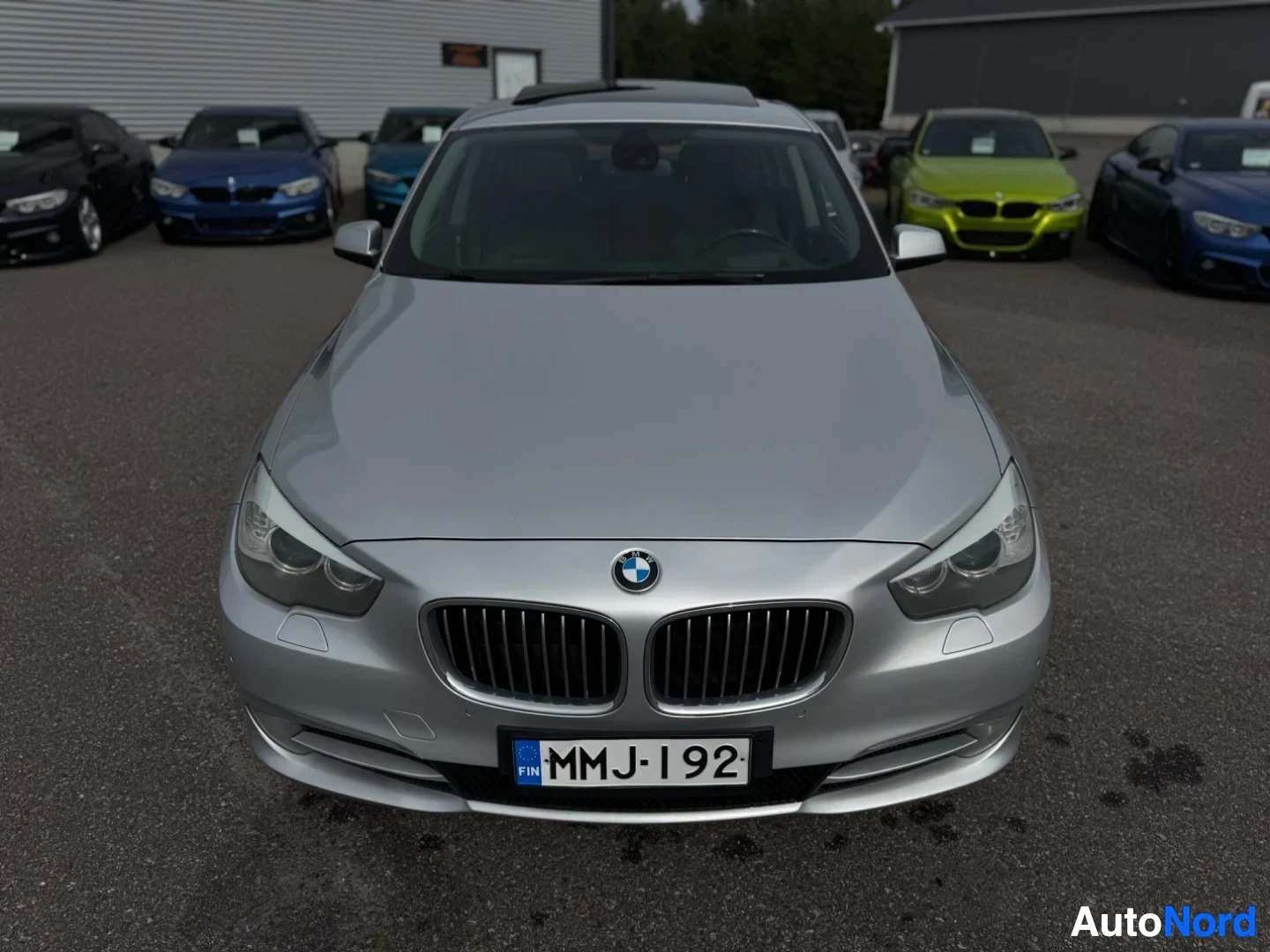 Edestä: Hopeanvärinen BMW edestäpäin kuvattuna. Autossa on musta etusäleikkö ja rekisterikilpi. Kattoluukku on näkyvissä. Taustalla näkyy rakennus ja muita auto