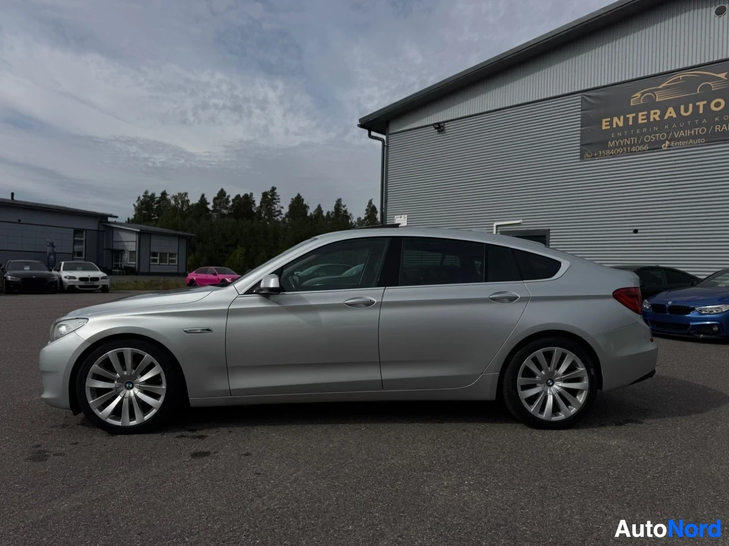 Sivulta: Hopeanvärinen henkilöauto oikealta puolelta. Auto on pysäköity asfaltille rakennuksen eteen, jossa lukee "ENTERAUTO".