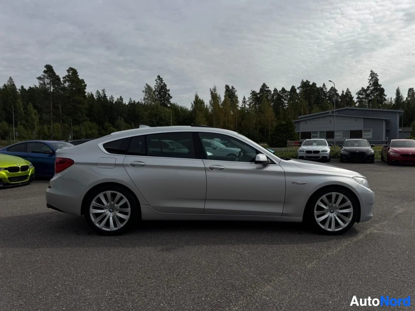 Sivulta: Hopeanvärinen henkilöauto vasemmalta puolelta. Auto on pysäköity asfaltille, taustalla puita ja rakennus.