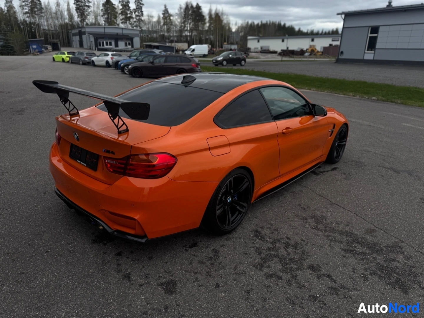 Takaviisto: Oranssi kaksiovinen coupé, jossa on iso musta takaspoileri, seisoo asfalttipihalla vasemmassa takaviistossa kuvattuna. Autossa on musta katto ja mustat vanteet.