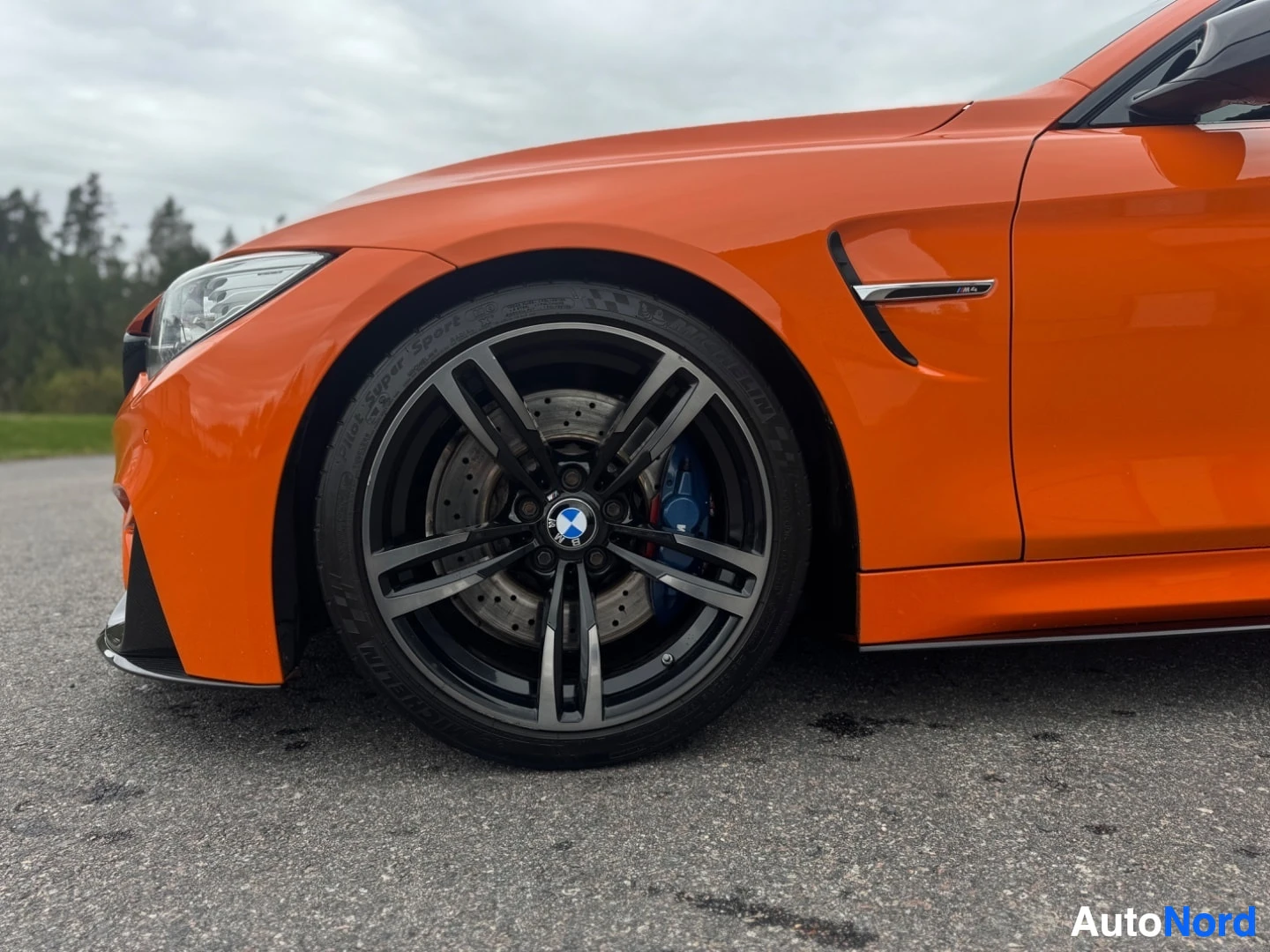 Yksityiskohta: Oranssin auton etupyörän ja lokasuojan lähikuva. Vanteet ovat mustat, ja siniset jarrusatulat näkyvät vanteen läpi. Renkaassa lukee "Michelin Pilot Super Sport"