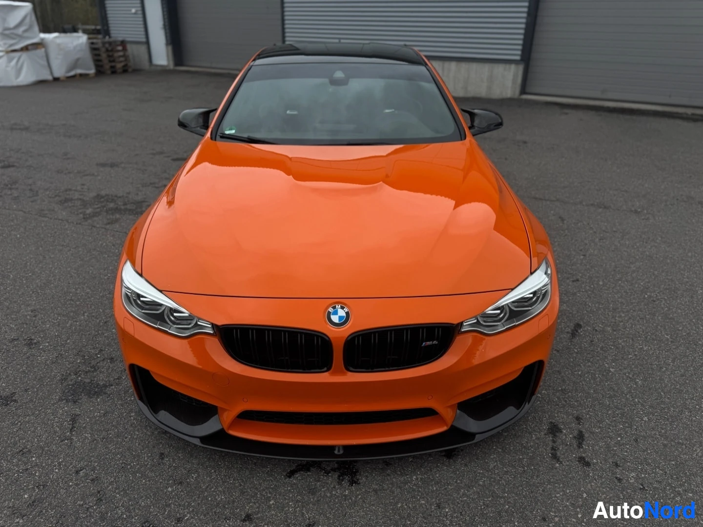Edestä: Oranssi kaksiovinen coupé, jossa on musta katto ja musta etusäleikkö. Auto on pysäköity ulos rakennuksen eteen ja kuvattu suoraan edestä ylhäältäpäin.