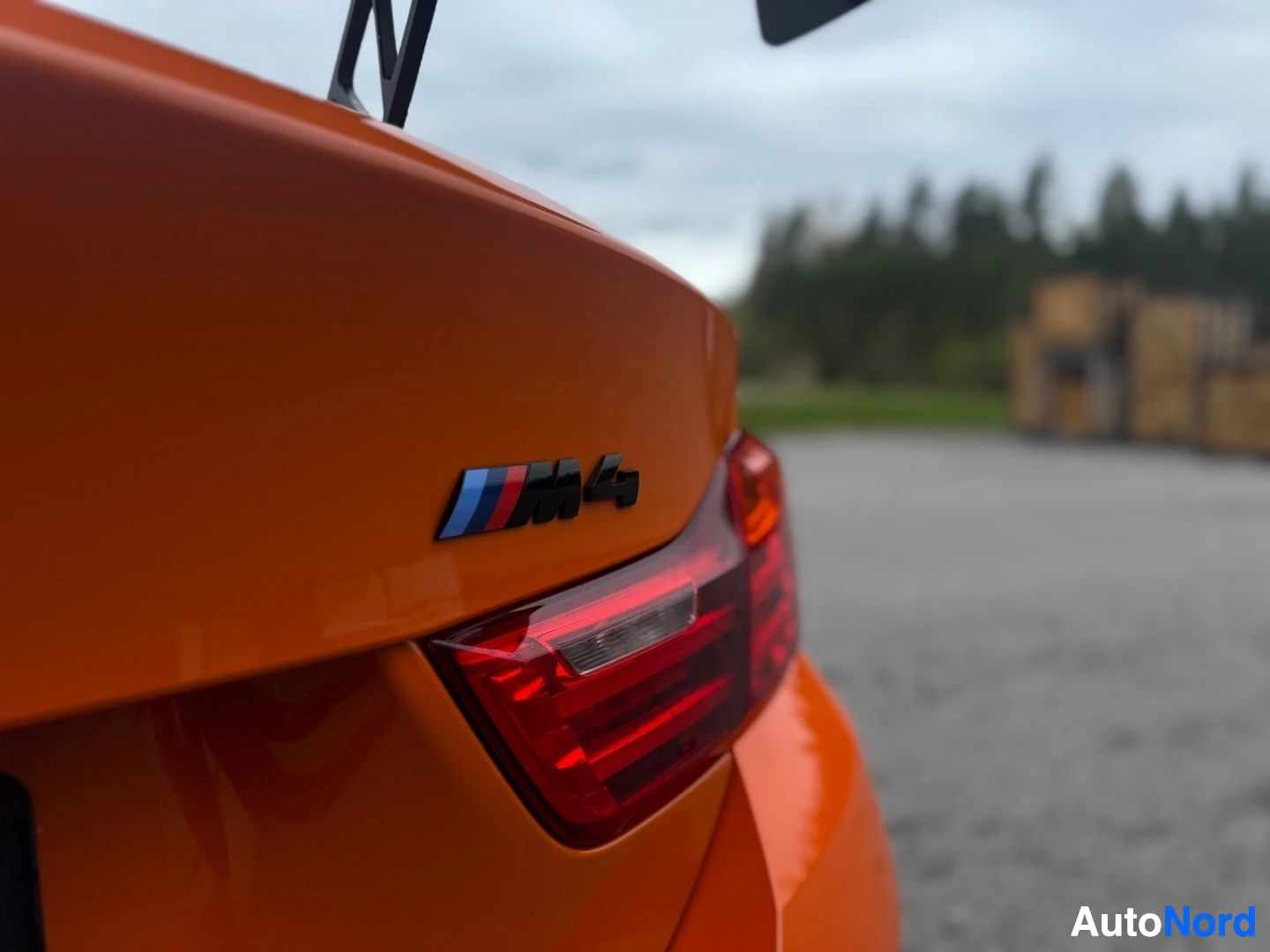 Yksityiskohta: Lähikuva auton oranssista takaluukusta, jossa on krominen ja sininen "M4"-logo. Oikealla näkyy osa takavalosta. Taustalla näkyy puita.