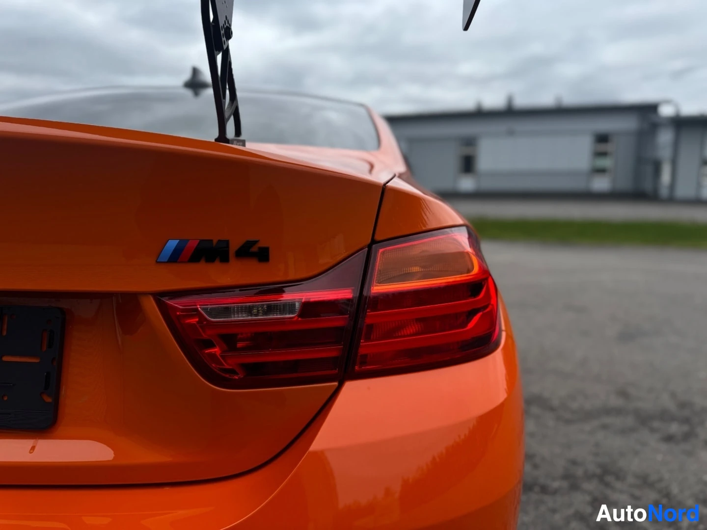 Takaviisto: Takakontin luukku, jossa näkyvät BMW M -logot ja M4-mallimerkintä, takavalo ja suuri takaspoileri. Auton väri on oranssi. Taustalla rakennuksia ja puita.