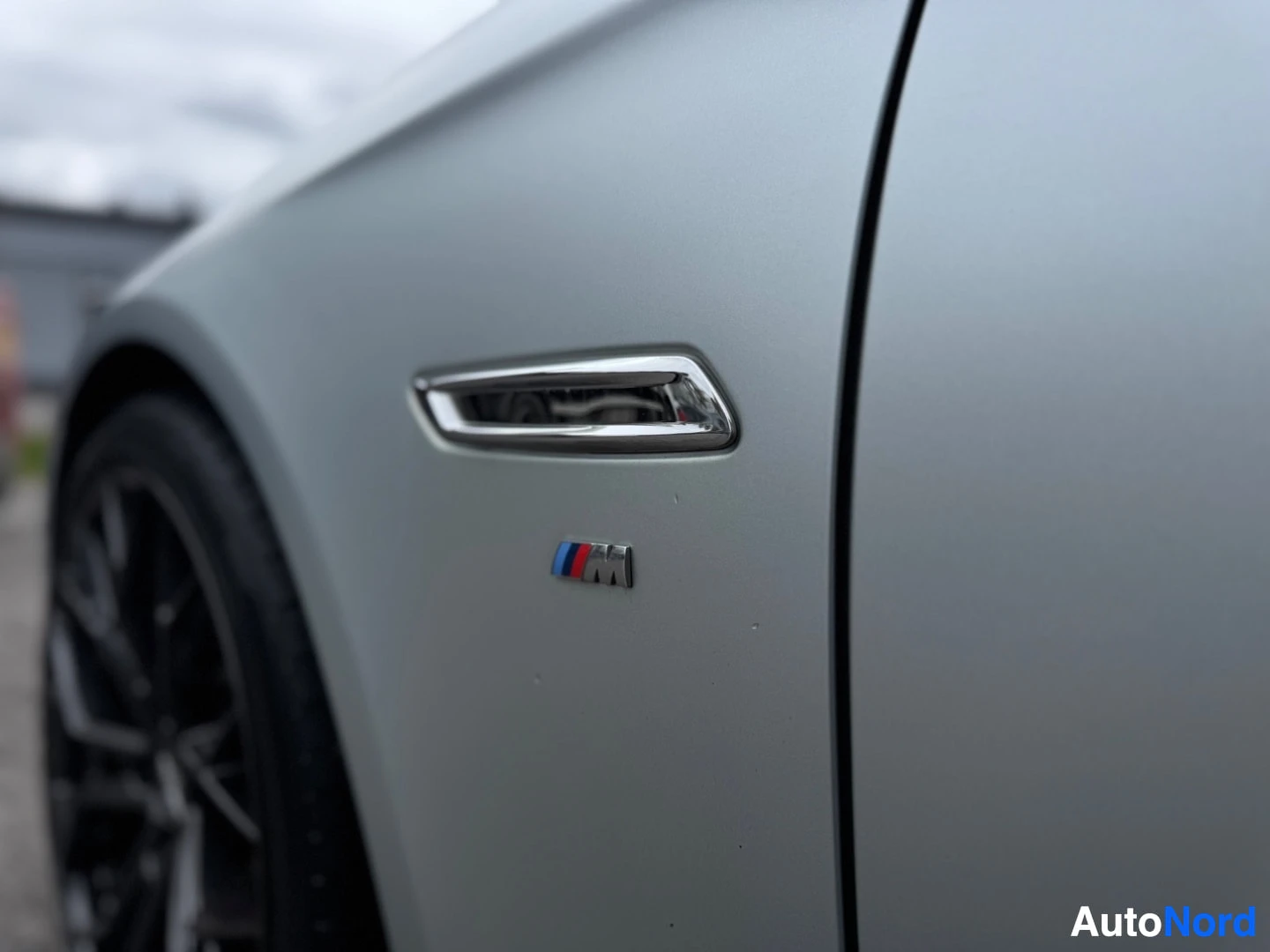 Sivulta: Lähikuva auton etulokasuojasta, jossa on pieni BMW M -logomerkki lokasuojan tuuletusaukon alapuolella.