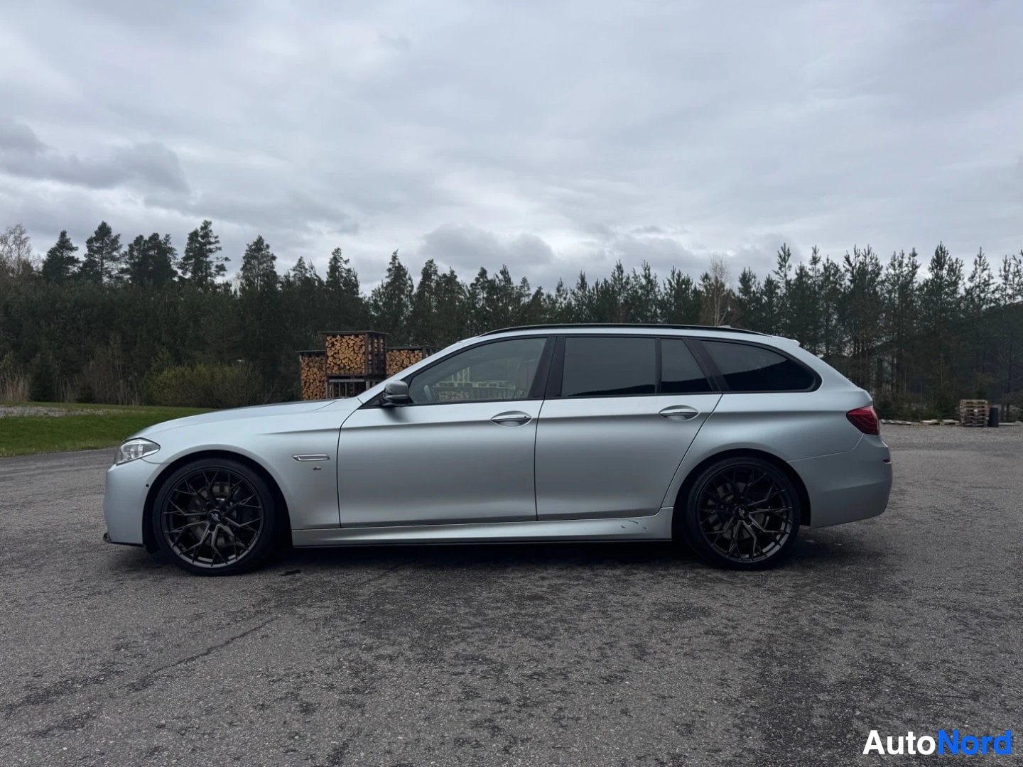 Sivulta: Hopeanvärinen farmariauto pysäköitynä ulos, vasen kylki näkyvissä. Autossa on mustat kevytmetallivanteet ja tummat takasivuikkunat. Taustalla näkyy puita.