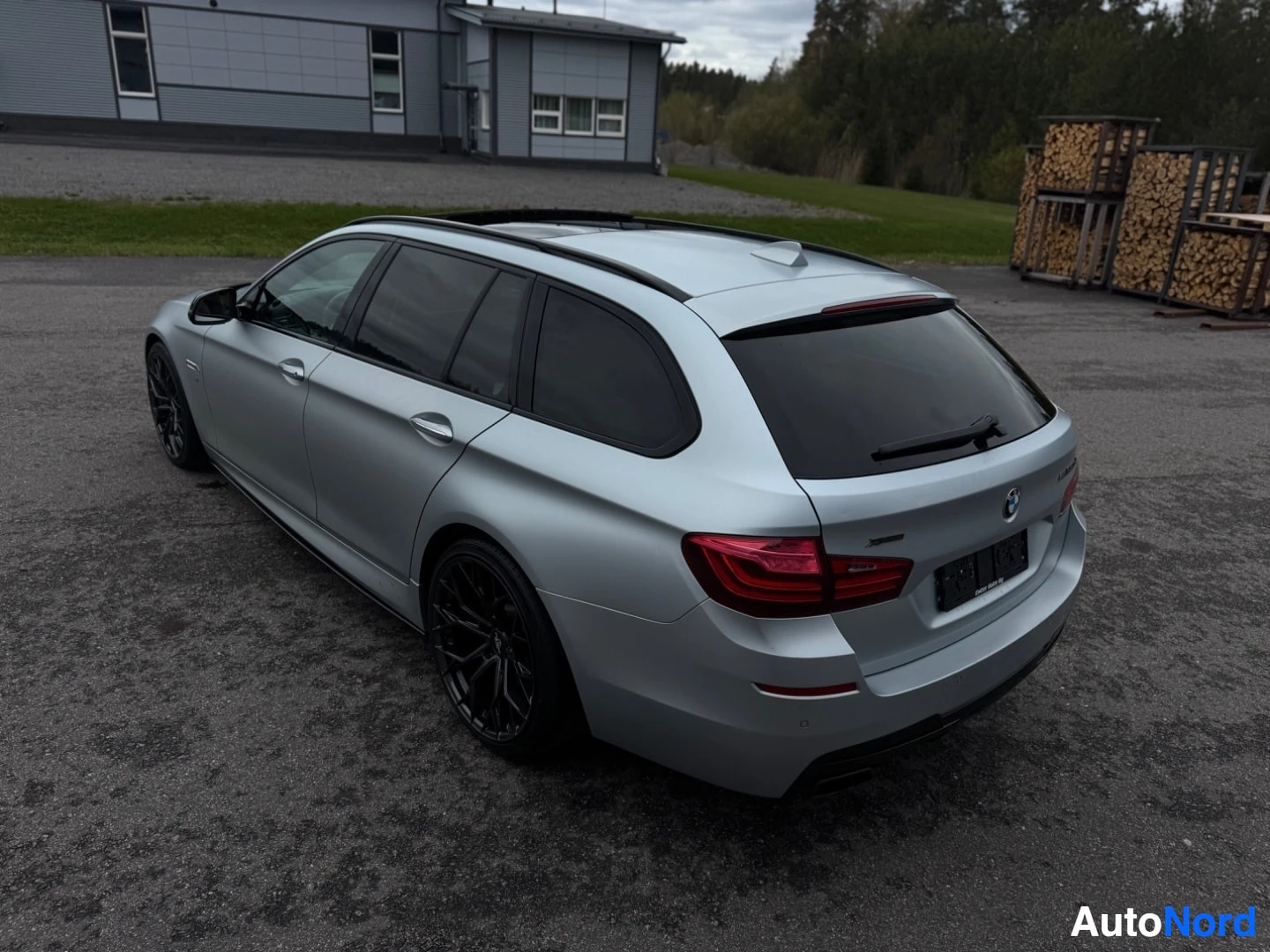 Takaviisto: Hopeanvärinen BMW 5-sarjan farmari pysäköitynä ulkona. Auto on kuvattu takaapäin yläviistosta vasemmalta ja näkyvissä ovat takapuskuri, takavalot, takaluukku, v