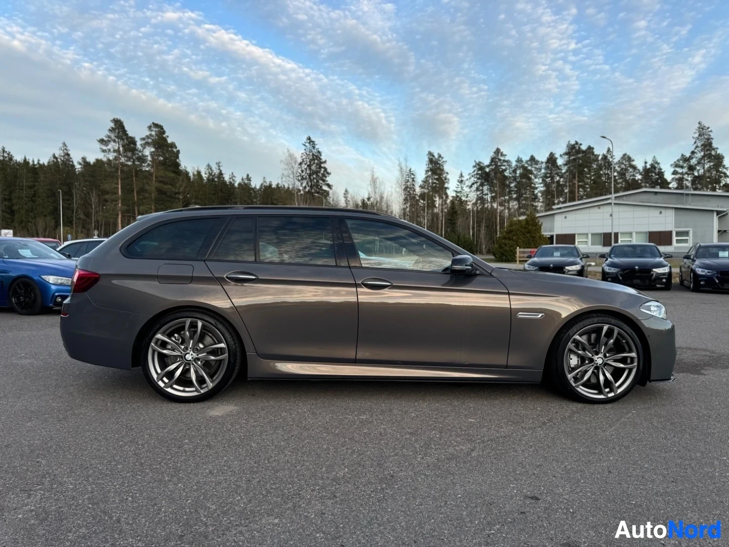 Sivulta: Harmaa farmariauto, jossa on kiiltävät monipuolaiset kevytmetallivanteet, pysäköity asfalttipihalle muiden ajoneuvojen ja puurivin eteen sinisen taivaan alla.