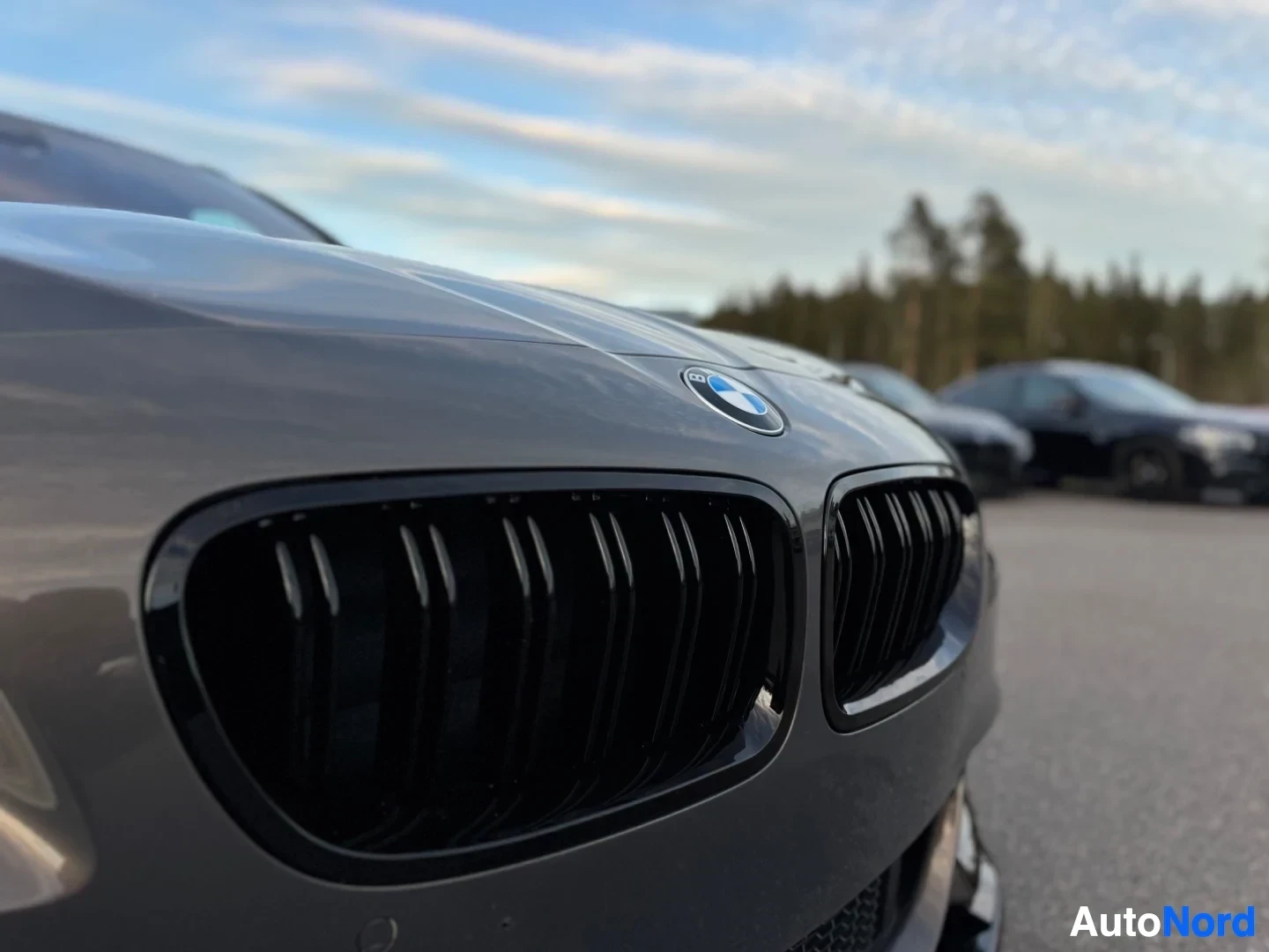 Etuviisto, nokka vasemmalle: Kuva auton etuosasta edestä ja hieman oikealta ylhäältä päin, keskittyen tummaan maskiin, jossa on vaakasuuntaiset rivat ja BMW-logo. Auton edessä ja k