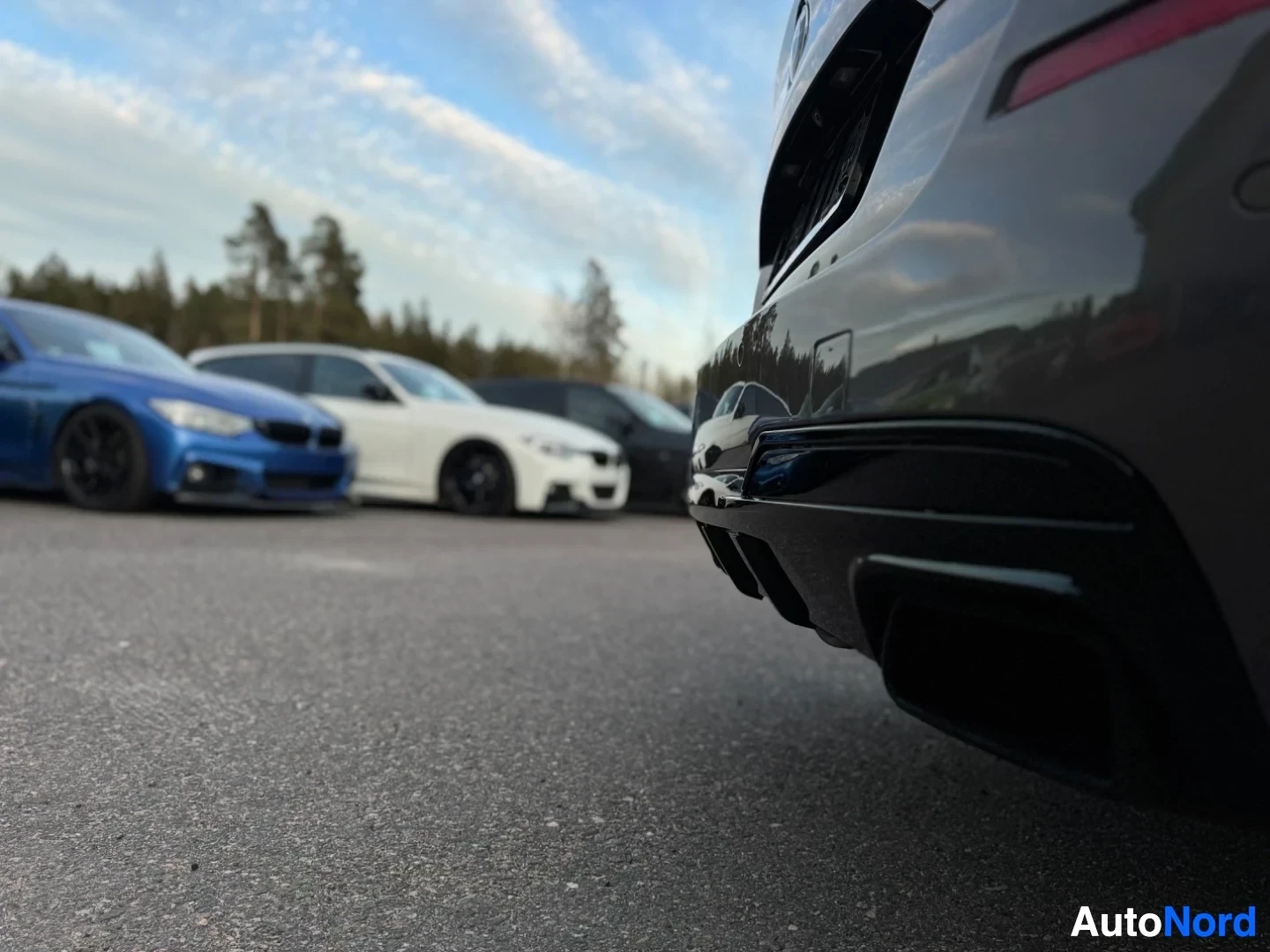Takaviisto: Lähikuva auton oikeasta takakulmasta, jossa näkyy pakoputkien päät, puskurin alaosa ja sumea tausta, jossa on muita autoja ja puita.