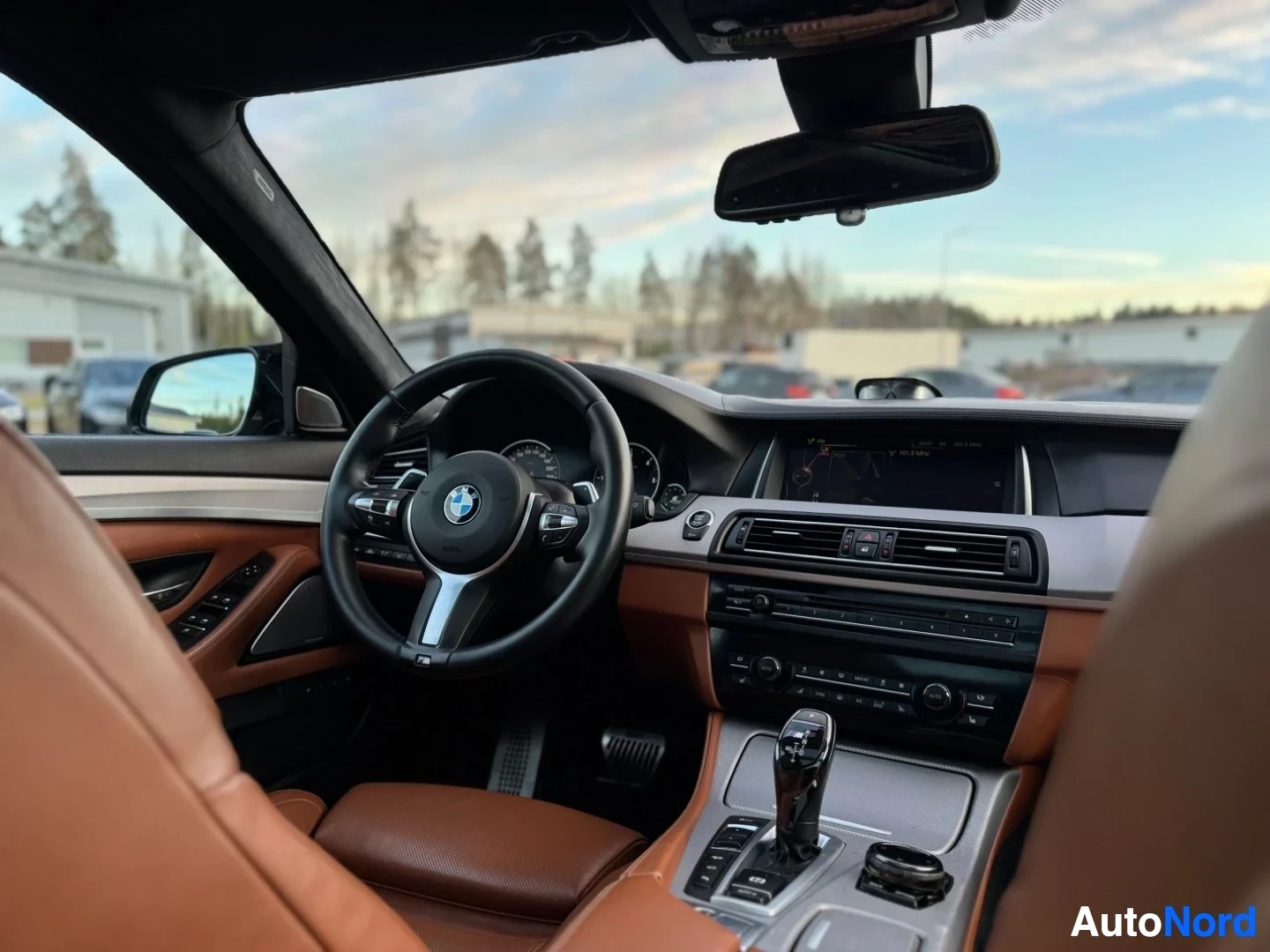 Ohjaamo: Sisäkuva auton etuosasta, jossa näkyvät ohjauspyörä, mittaristo ja infotainment-näyttö. Ruskeat istuimet ja hopeanväriset koristelistat näkyvät myös, taustalla