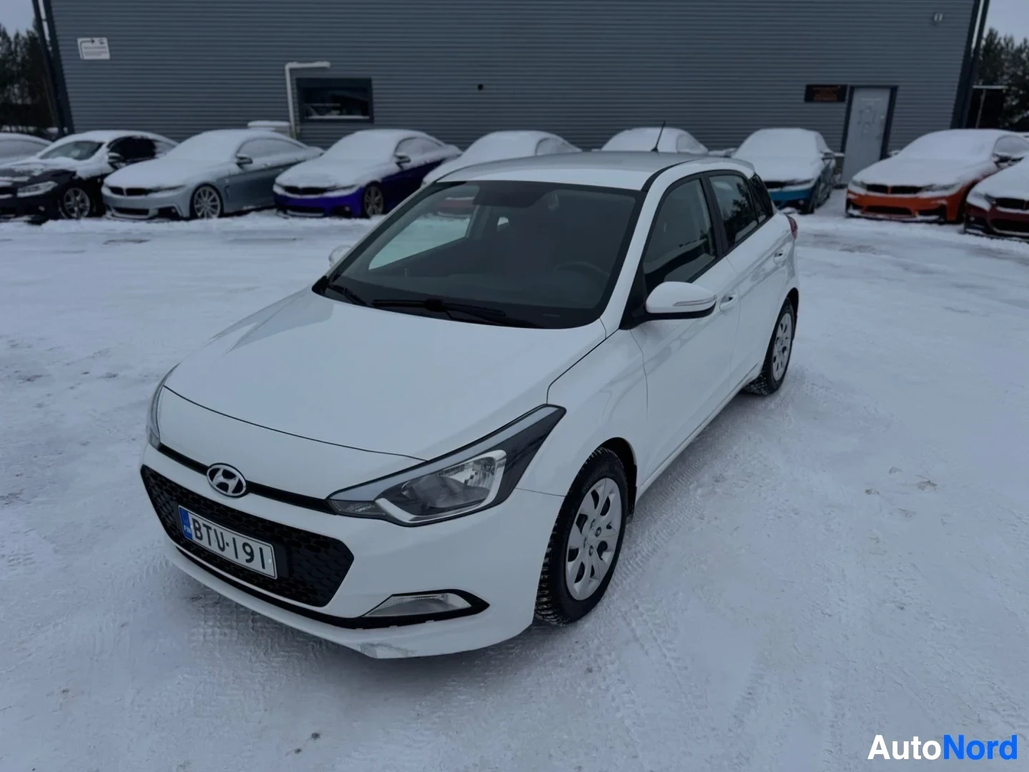 Etuviisto, nokka vasemmalle: Valkoinen Hyundai i20 -henkilöauto pysäköitynä lumiselle piha-alueelle. Autossa on mustat monipuolaiset vanteet ja rekisterikilpi "" edessä. Tau