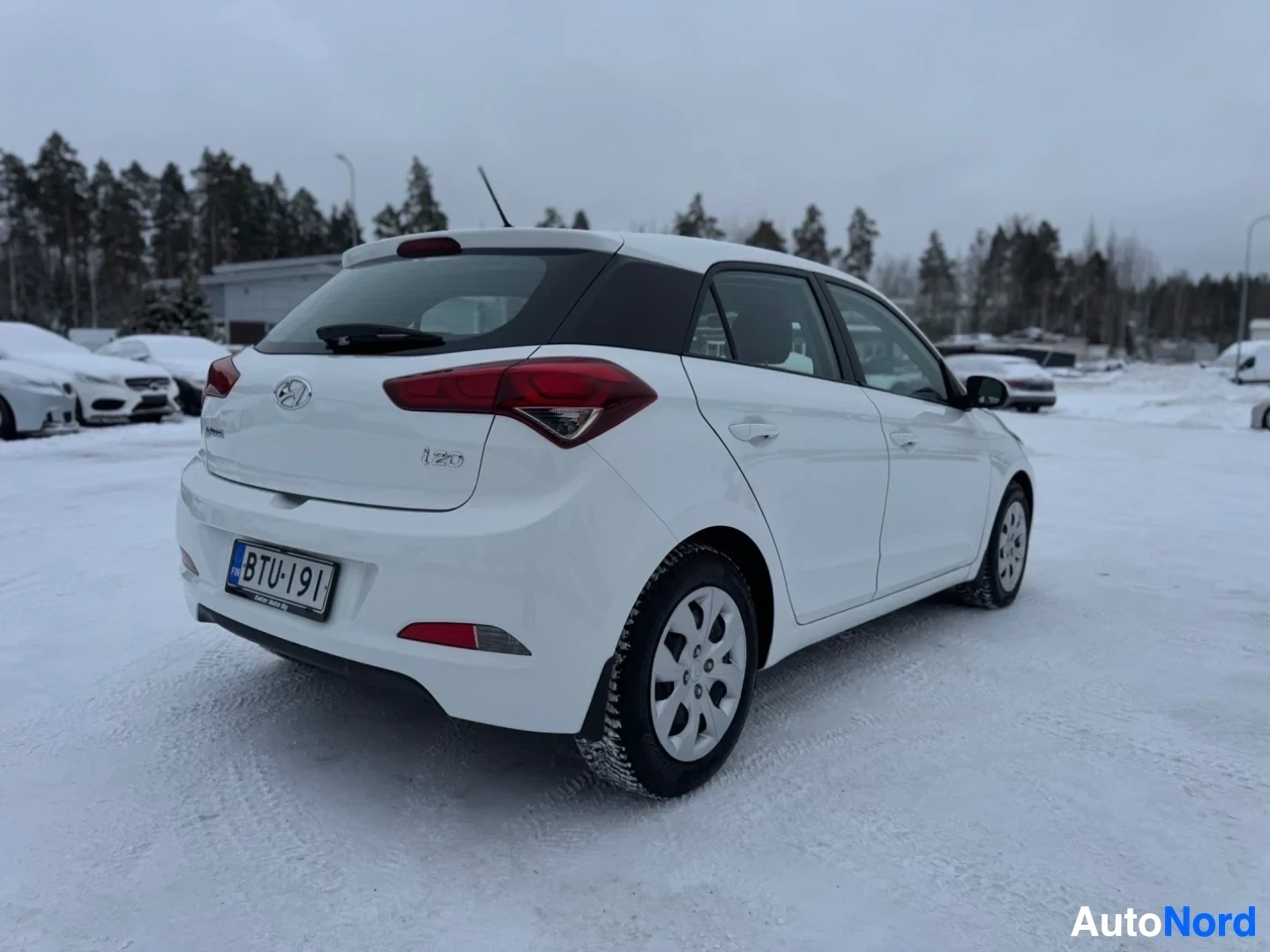 Takaviisto: Valkoinen viistoperäinen henkilöauto on pysäköity lumiselle kentälle. Ajoneuvon vasen takakulma on näkyvissä. Rekisterikilvessä lukee