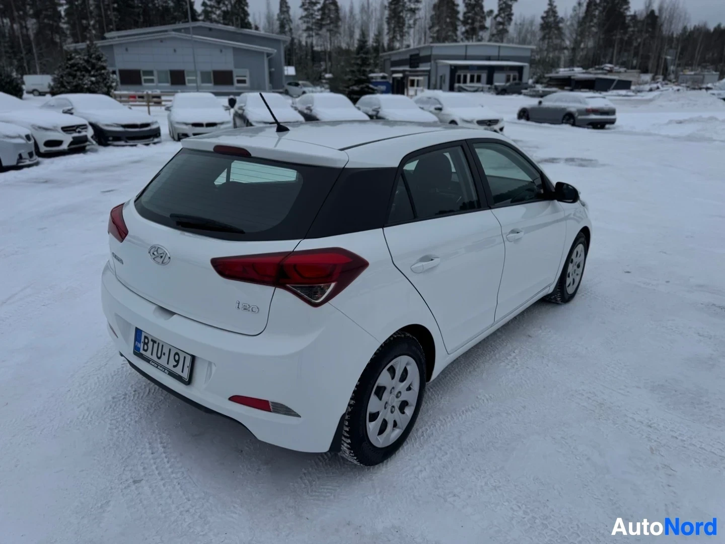Takaviisto: Valkoinen viistoperäinen henkilöauto on pysäköity lumiselle alueelle. Ajoneuvon vasen takaosa on kuvassa. Rekisterikilpi on näkyvillä ja siinä lukee