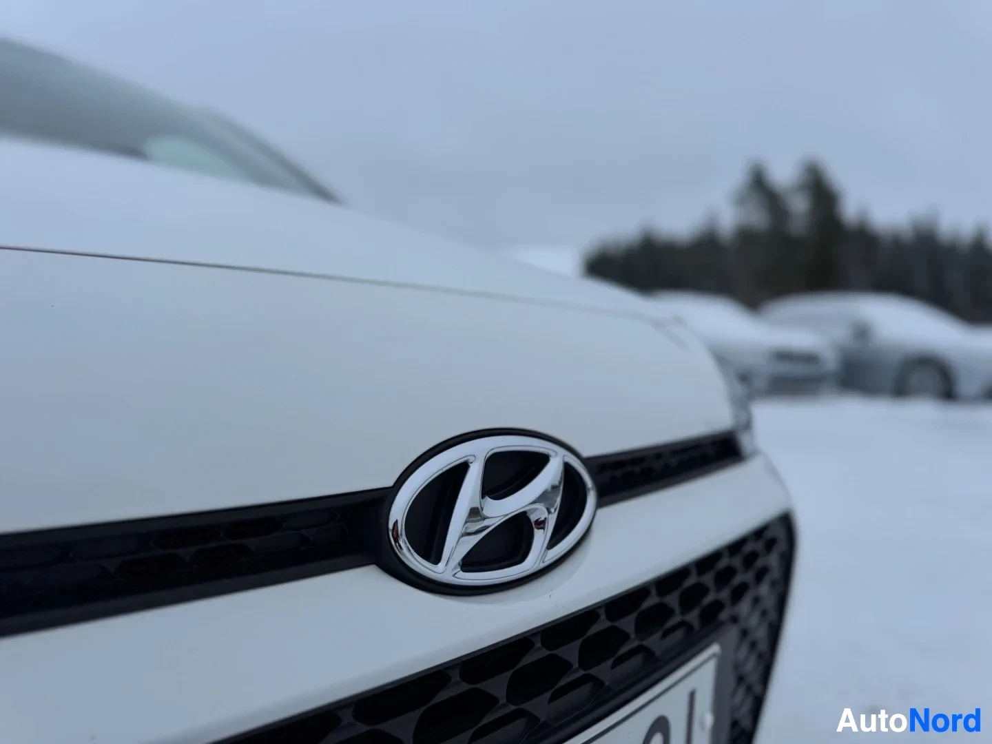 Yksityiskohta: Lähikuva auton etusäleiköstä, jossa on kromattu Hyundai-logo. Auto on lumisella parkkipaikalla, ja taustalla näkyy epäselviä puita.