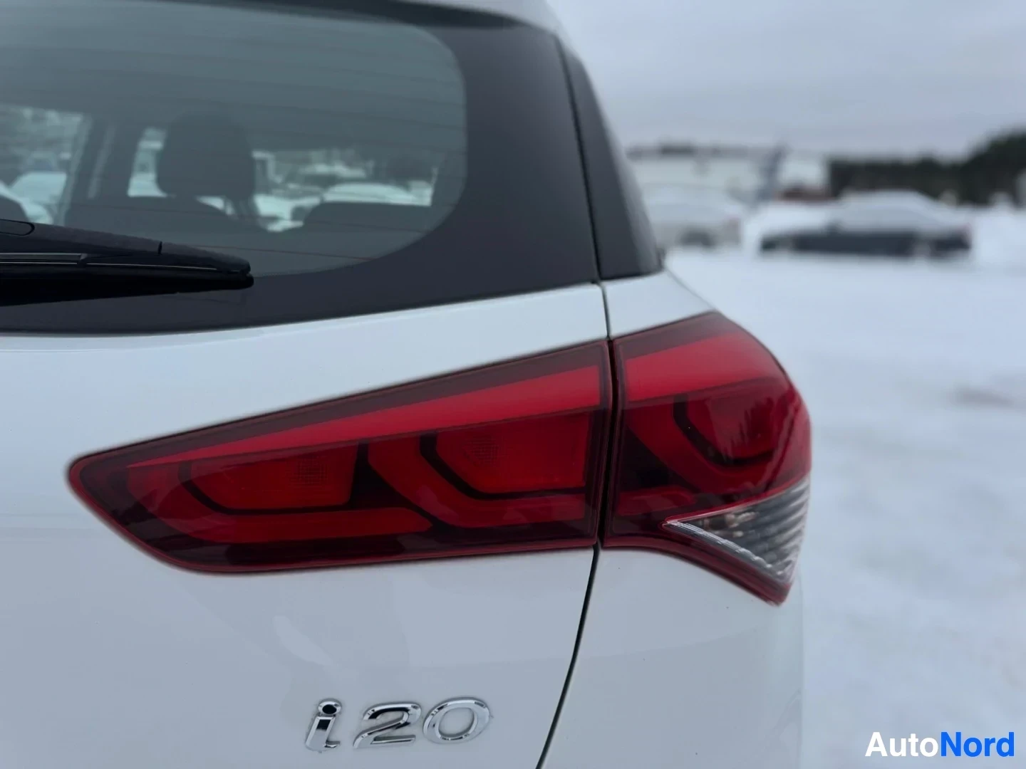 Takaviisto: Lähikuva valkoisen auton takaosasta, jossa näkyy tummanpunainen takavalo ja takalasinpyyhin. Lumipeitteinen ympäristö ja muita autoja näkyvät taustalla sumeana.