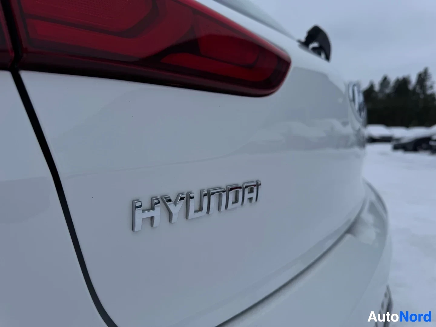 Takaviisto: Lähikuva valkoisen auton takaluukusta, jossa on krominvärinen "HYUNDAI"-logo. Taustalla näkyy lumista maata ja epäselviä puita.