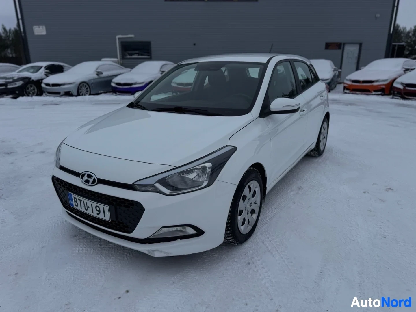 Etuviisto, nokka vasemmalle: Valkoinen Hyundai i20 -henkilöauto pysäköitynä lumiselle piha-alueelle harmaan rakennuksen eteen. Ajoneuvossa on mustat, useampipuolaiset vanteet ja et