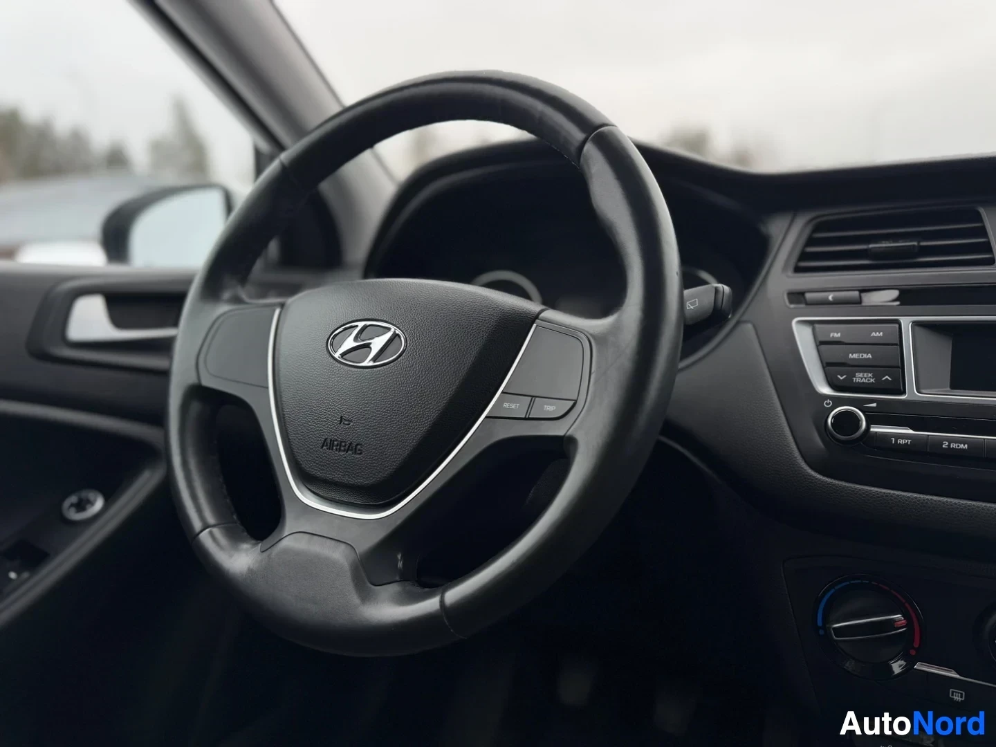 Yksityiskohta: Lähikuva auton ohjauspyörästä, jossa on Hyundai-logo ja airbag-teksti keskellä. Ohjauspyörän oikealla puolella on kolme painiketta ja vasemmalla puolella näkyy