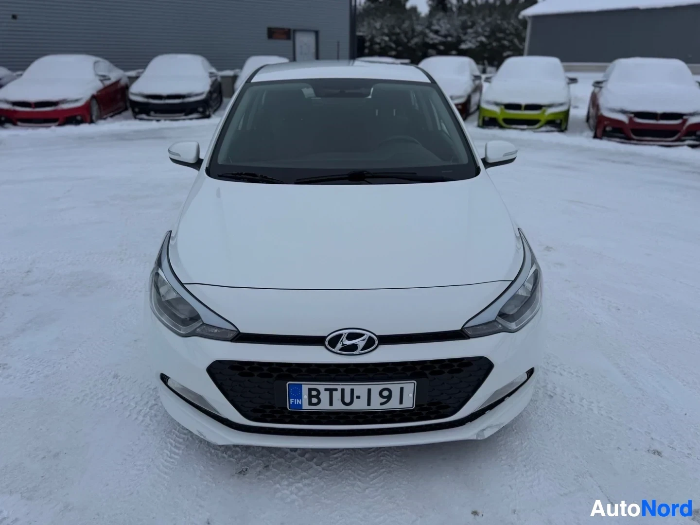 Edestä: Valkoinen Hyundai i20 -henkilöauto edestä päin lumisella maalla. Autossa on musta hunajakennosäleikkö, sumuvalot edessä ja rekisterikilpi ""