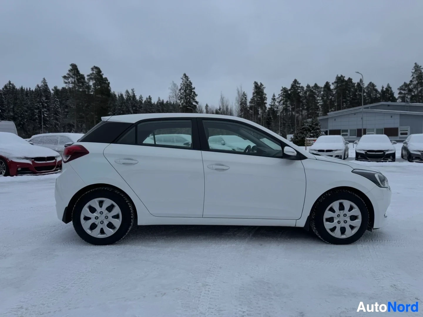 Sivulta: Valkoinen henkilöauto lumisella parkkipaikalla, kuvattuna suoraan sivusta. Autossa on mustat ikkunakehykset ja takaspoileri.