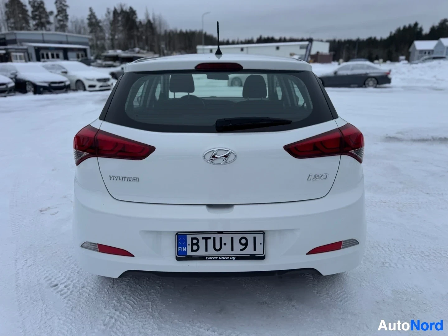 Takaa: Valkoinen viistoperäinen henkilöauto lumisella parkkipaikalla, kuvattuna suoraan takaa. Auton takaluukku, takavalot ja rekisterikilpi ovat selkeästi näkyvillä.