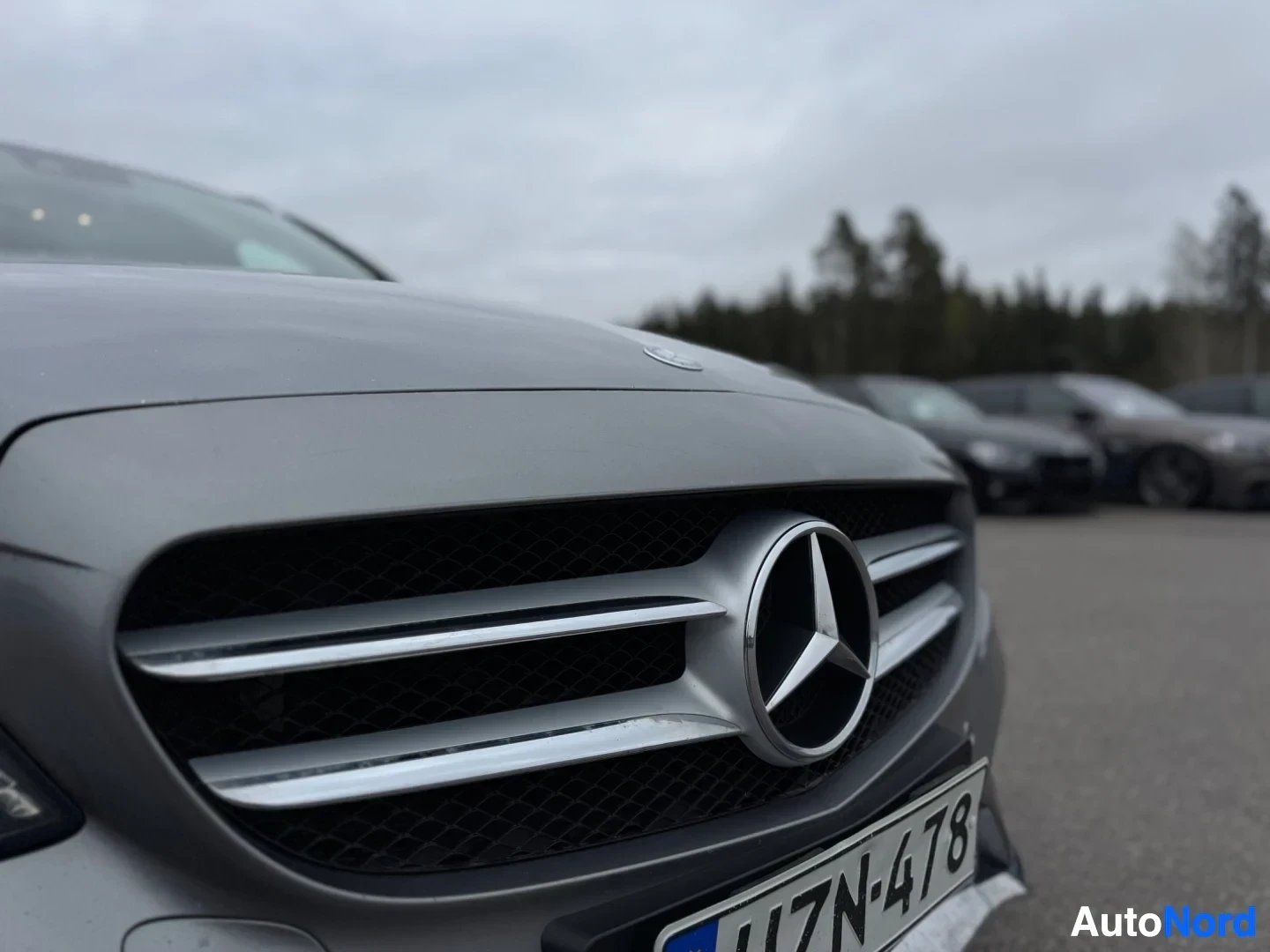 Yksityiskohta: Lähikuva harmaan henkilöauton keulasta. Keulagrilli ja Mercedes-Benzin tähtilogo ovat selkeästi näkyvillä. Rekisterikilven alareuna on osittain näkyvissä, kilve
