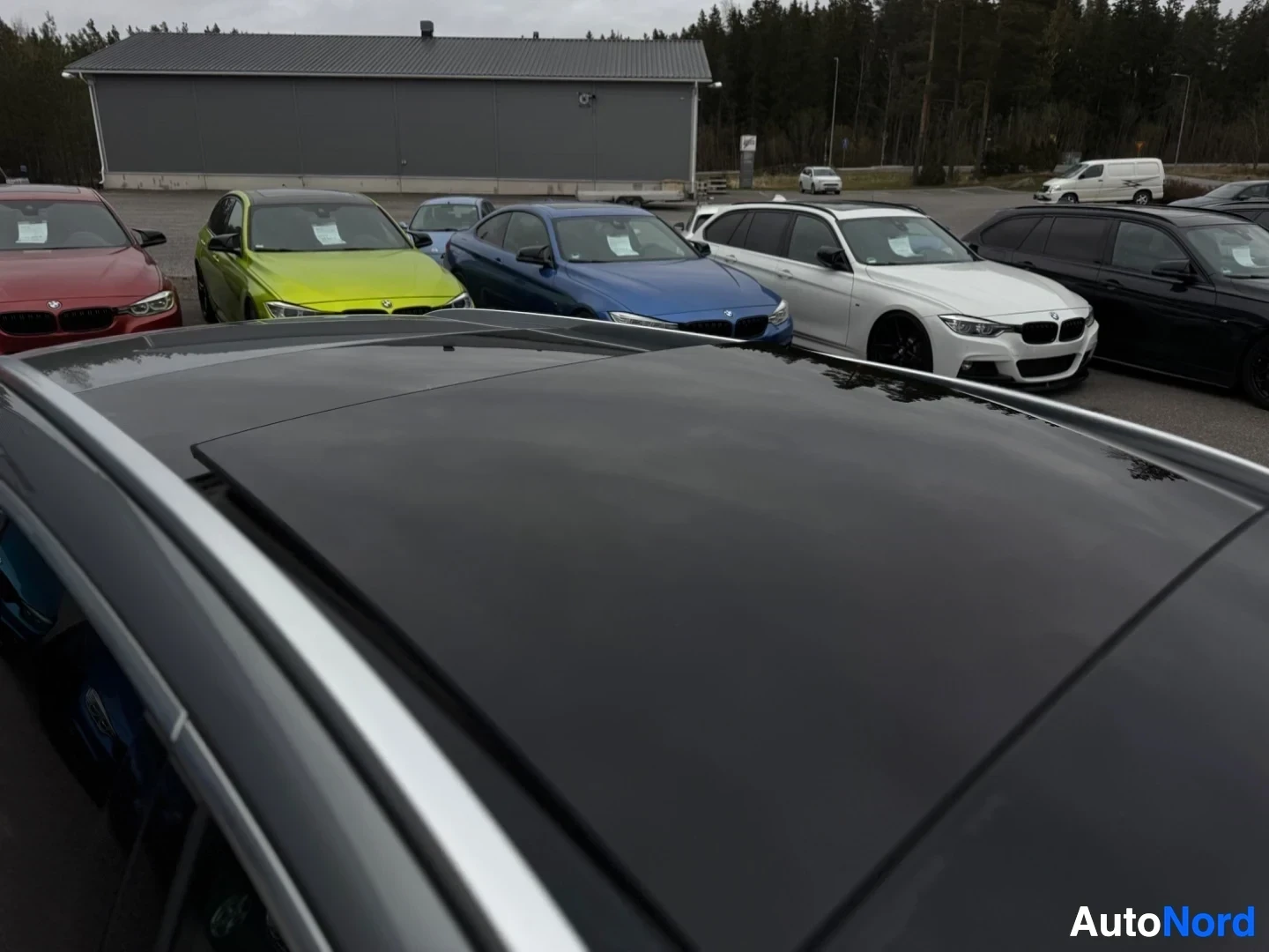 Yksityiskohta: Auton harmaa panoraamalasikatto kuvattuna ylhäältä ja takaa viistosti. Taustalla näkyy useita pysäköityjä henkilöautoja ja rakennus.
