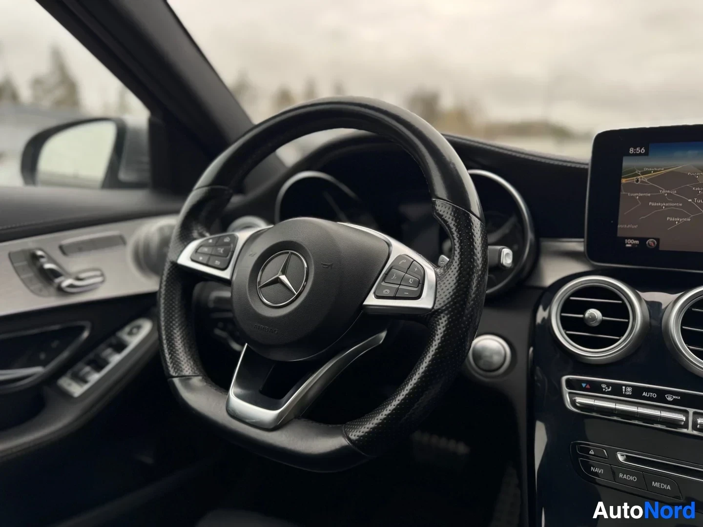 Yksityiskohta: Lähikuva auton ohjauspyörästä, jossa on Mercedes-Benz-logo keskellä ja monitoimipainikkeet puolissa. Kojelautanäytöllä näkyy navigaatio. Oikealla alhaalla näkyy