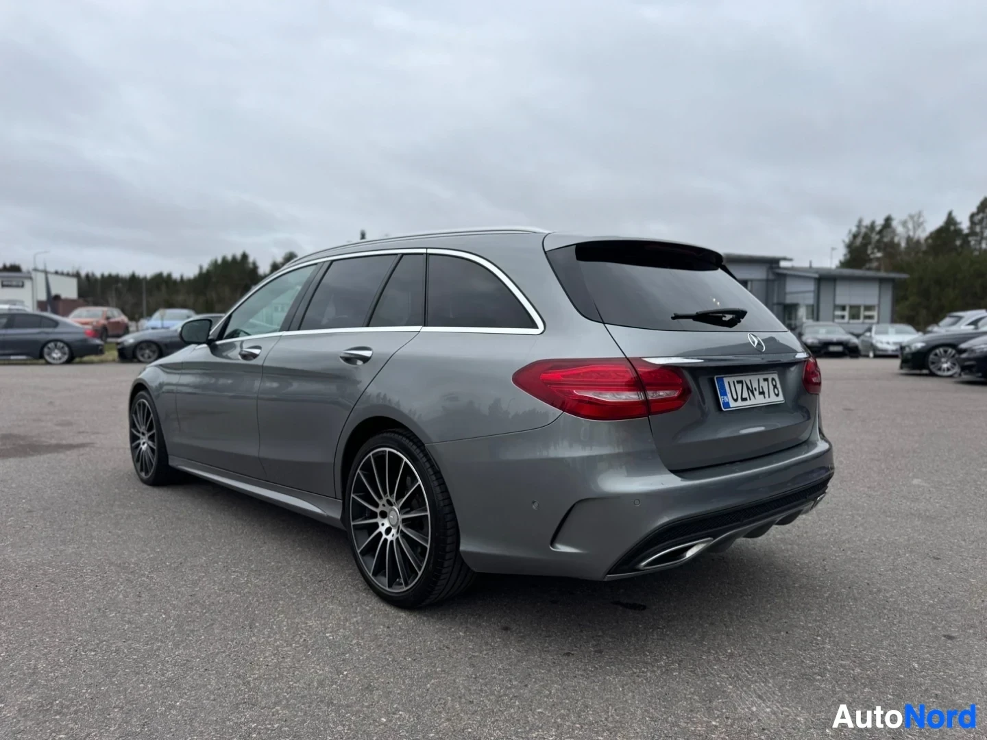Takaviisto: Harmaa farmariauto takaa oikealta puolelta kuvattuna pysäköintialueella. Autossa on mustat monipuolaiset kevytmetallivanteet ja kattokaiteet. Takana rekisteriki