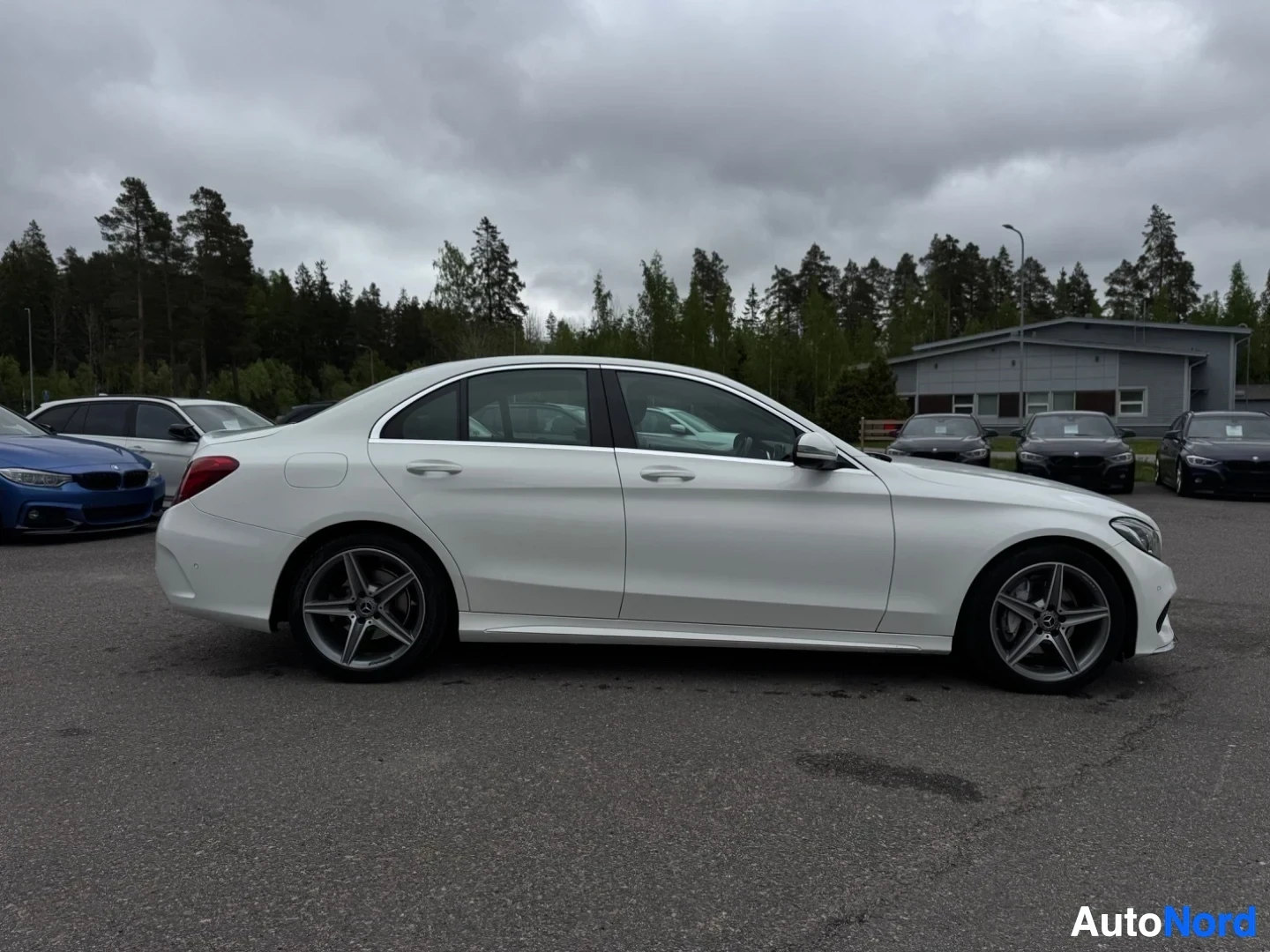 Sivulta: Valkoinen Mercedes-Benz C-sarjan sedan oikealta sivulta kuvattuna. Autossa on monipuoliset alumiinivanteet. Taustalla on muita pysäköityjä autoja ja metsäinen a