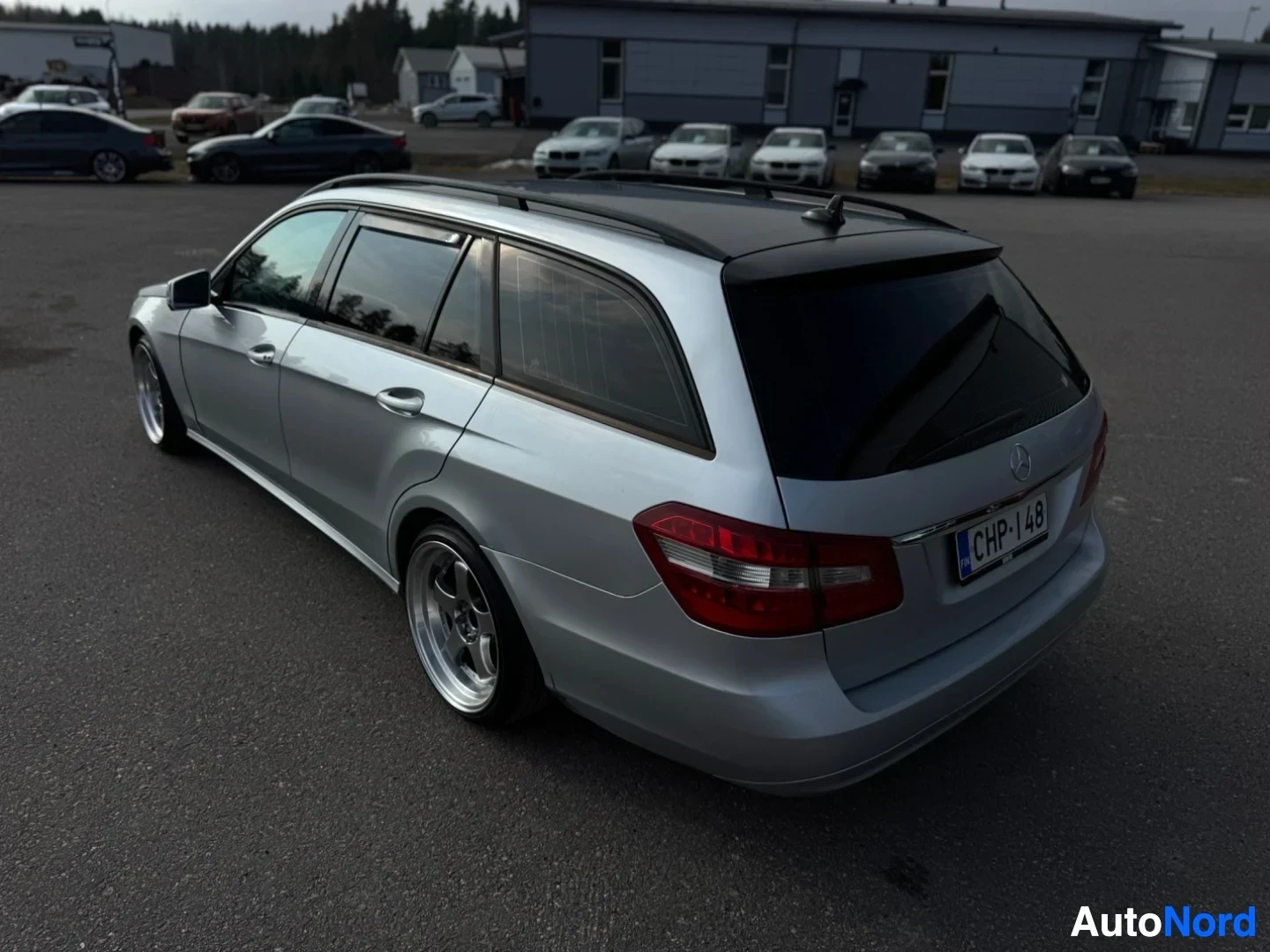 Takaviisto: Hopeanvärinen Mercedes-Benz E-sarjan farmariauto on pysäköity asfaltille takaosa vinosti kuvattuna. Autossa on kattokaiteet ja tumma panoraamakatto. Ajoneuvon t
