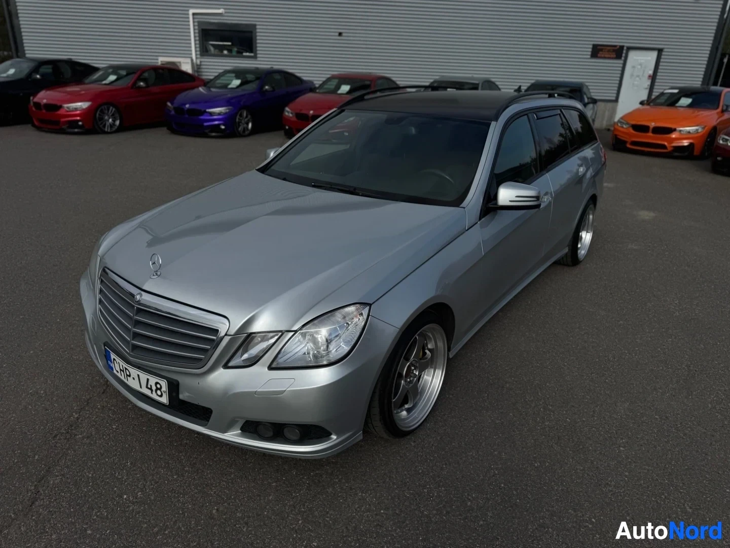 Etuviisto, nokka vasemmalle: Hopeanvärinen Mercedes-Benz E-sarjan farmari, jossa on tumma katto ja monipuolaiset kevytmetallivanteet, pysäköitynä myyntiliikkeen eteen.