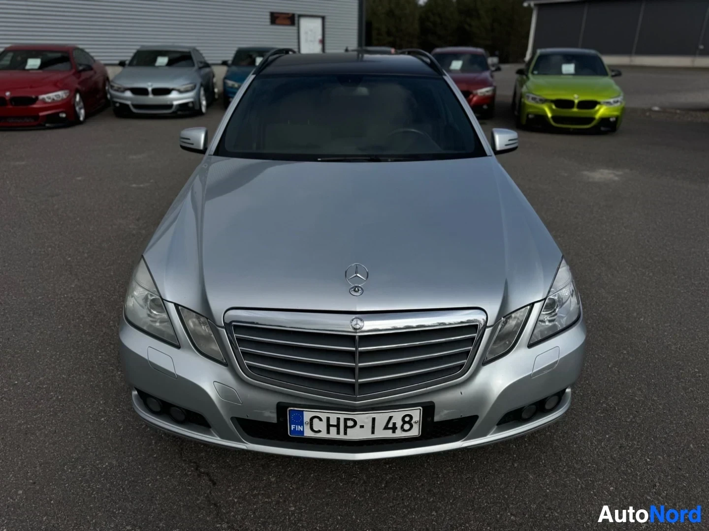 Edestä: Edestä kuvattu hopeanvärinen Mercedes-Benz E-sarjan farmari, jossa on tumma katto. Auto on pysäköity pihalle muiden autojen eteen.