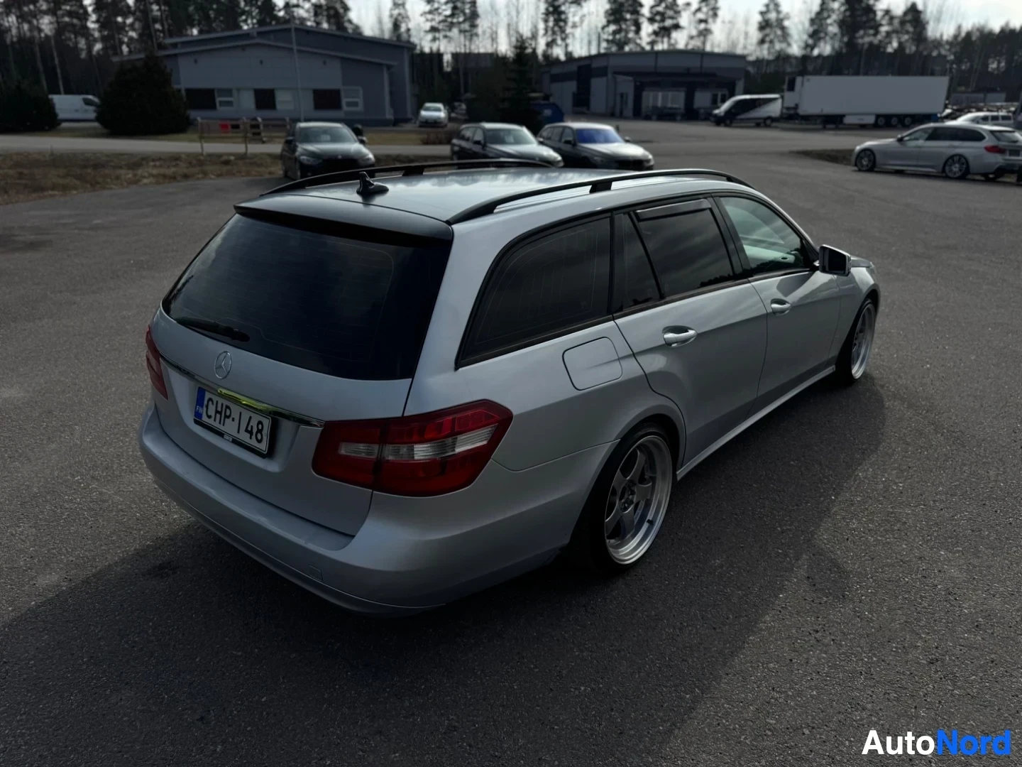 Takaviisto: Hopeanvärinen farmariauto takaoikealta kuvattuna ulkona pysäköintialueella. Autossa on kattokiskot ja tummennetut takalasit. Rekisterikilpi on näkyvissä