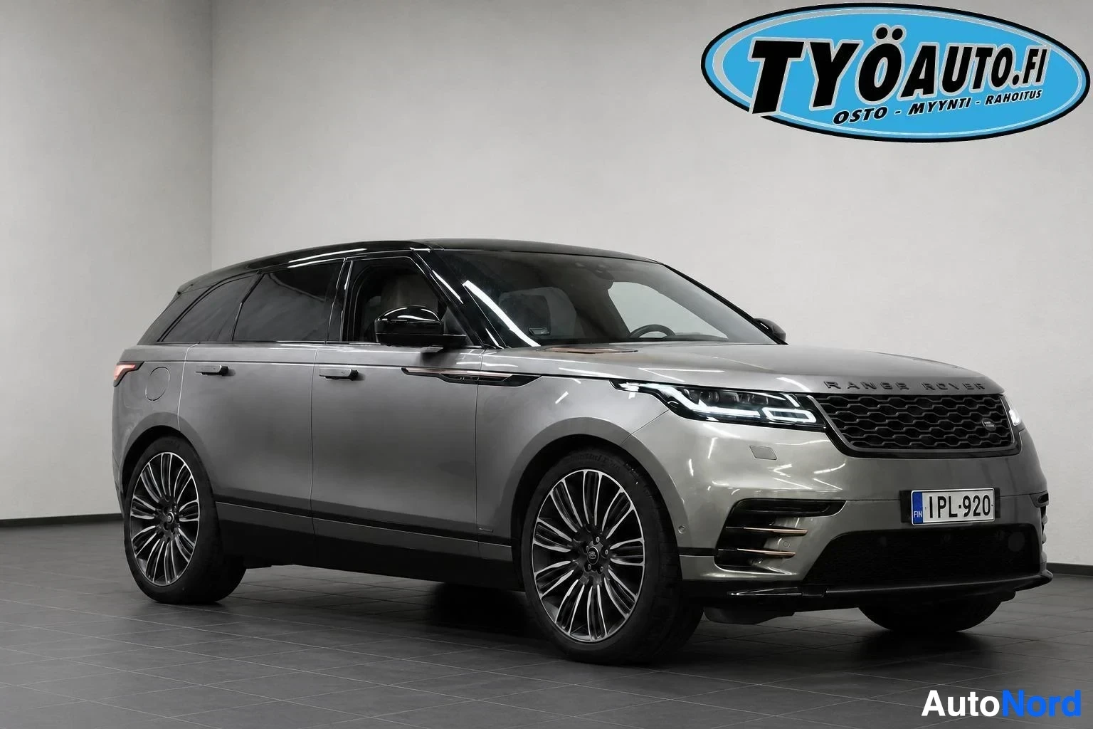 Etuviisto, nokka vasemmalle: Harmaa Range Rover Velar edestä ja oikealta sivulta kuvattuna autohallissa. Autossa on musta katto ja mustat monipuolaiset kevytmetallivanteet. Rekiste