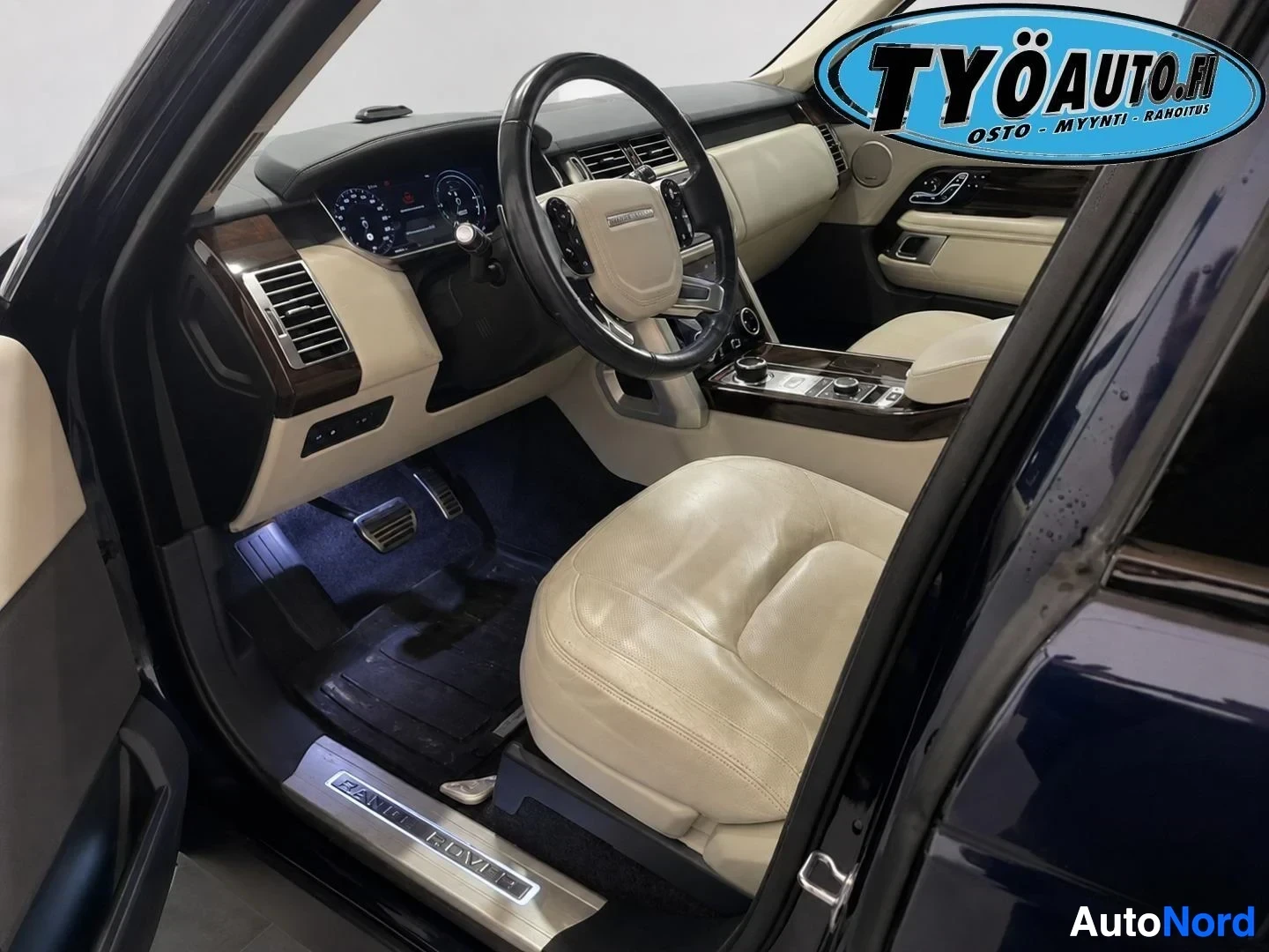 Ohjaamo: Kuljettajan puolen ovi on auki, ja sisältä näkyy vaaleanbeige nahkaverhoiltu istuin, ohjauspyörä ja kojelauta. Kynnyslevyssä lukee "RANGE ROVER".