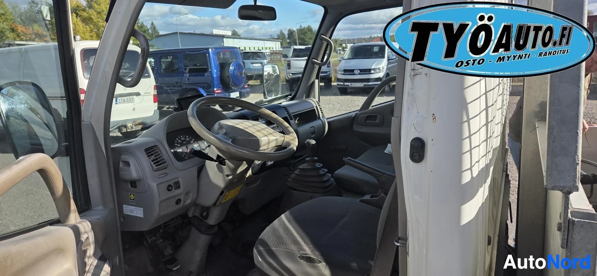 Ohjaamo: Kuva kuorma-auton ohjaamosta, jossa näkyy kolmipuolainen ohjauspyörä, mittaristo ja käsivalintainen vaihdekeppi lattialla. Kaksi istuinta näkyvissä.