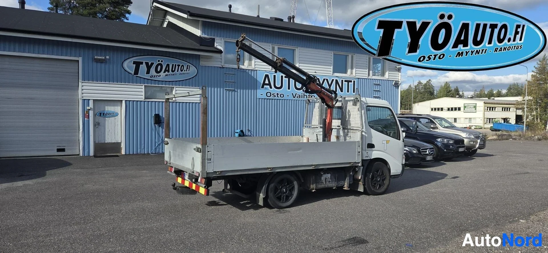 Takaviisto: Kuorma-auto nosturilla, autossa on valkoiset kori- ja ovipaneelit ja konepelti. Auto seisoo asfalttipihalla rakennuksen edessä. Nosturi on auton lavalla.