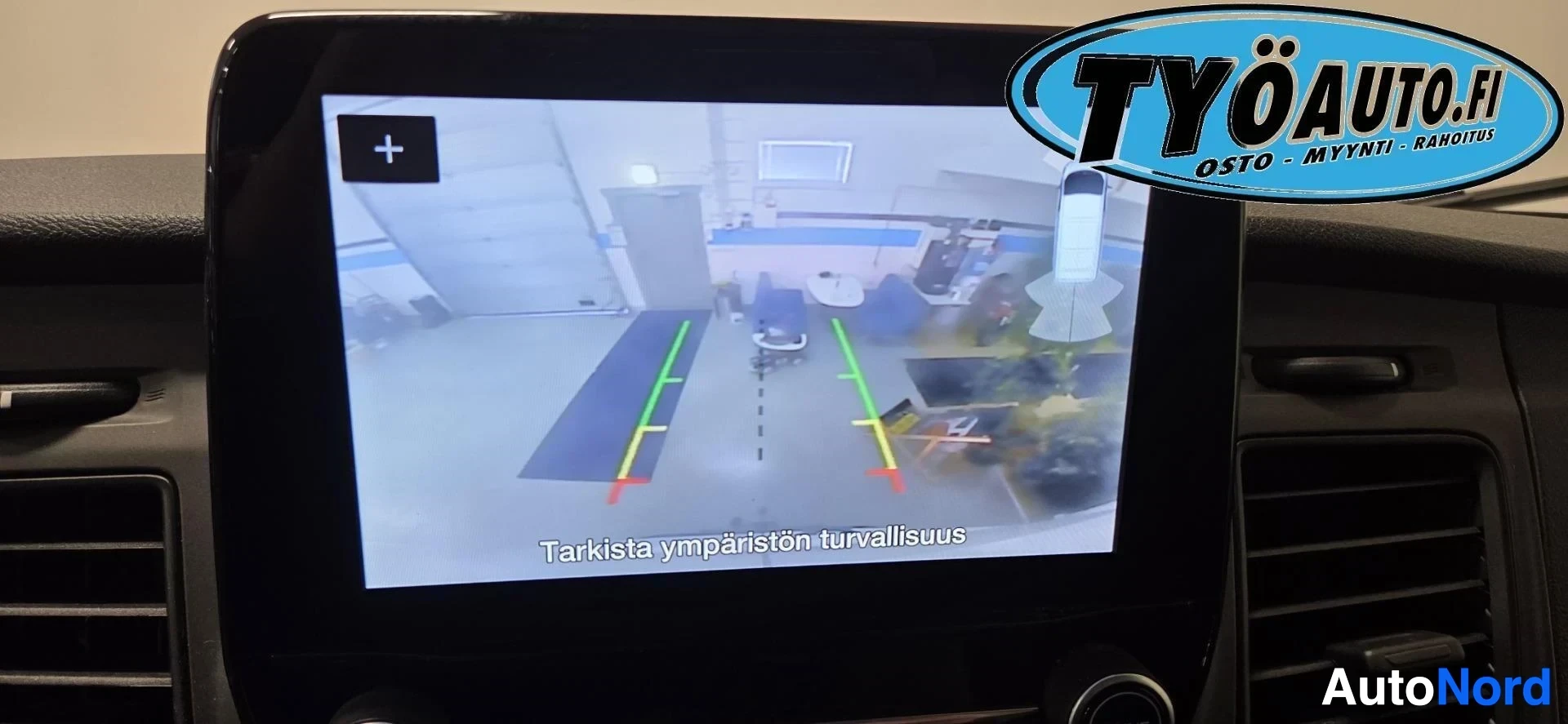Yksityiskohta: Lähikuva auton infotainment-näytöstä, jossa näkyy peruutuskameran kuva opastusviivoineen. Ruudulla lukee "Tarkista ympäristön turvallisuus". Kameranäkymässä näk