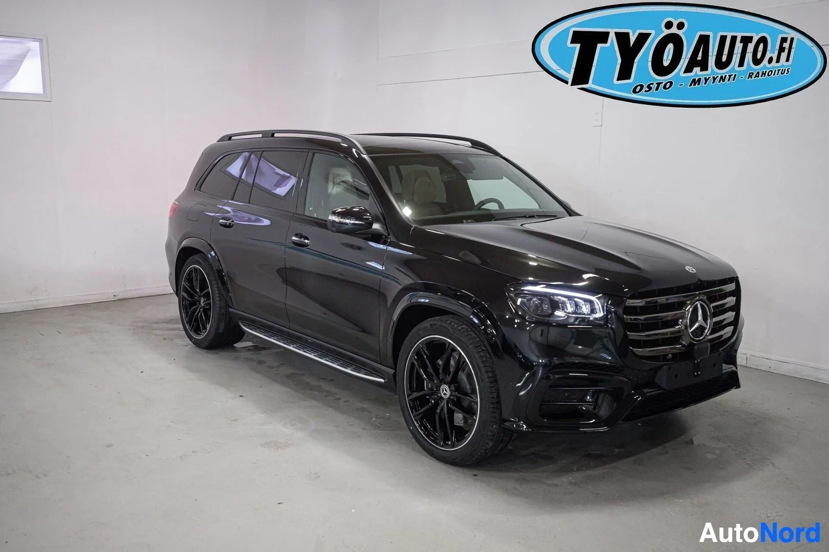 Etuviisto, nokka oikealle: Musta Mercedes-Benz GLS SUV edestä ja oikealta sivulta kuvattuna valkoisessa hallissa. Autossa on mustat monipuolaiset vanteet ja kattokaiteet.