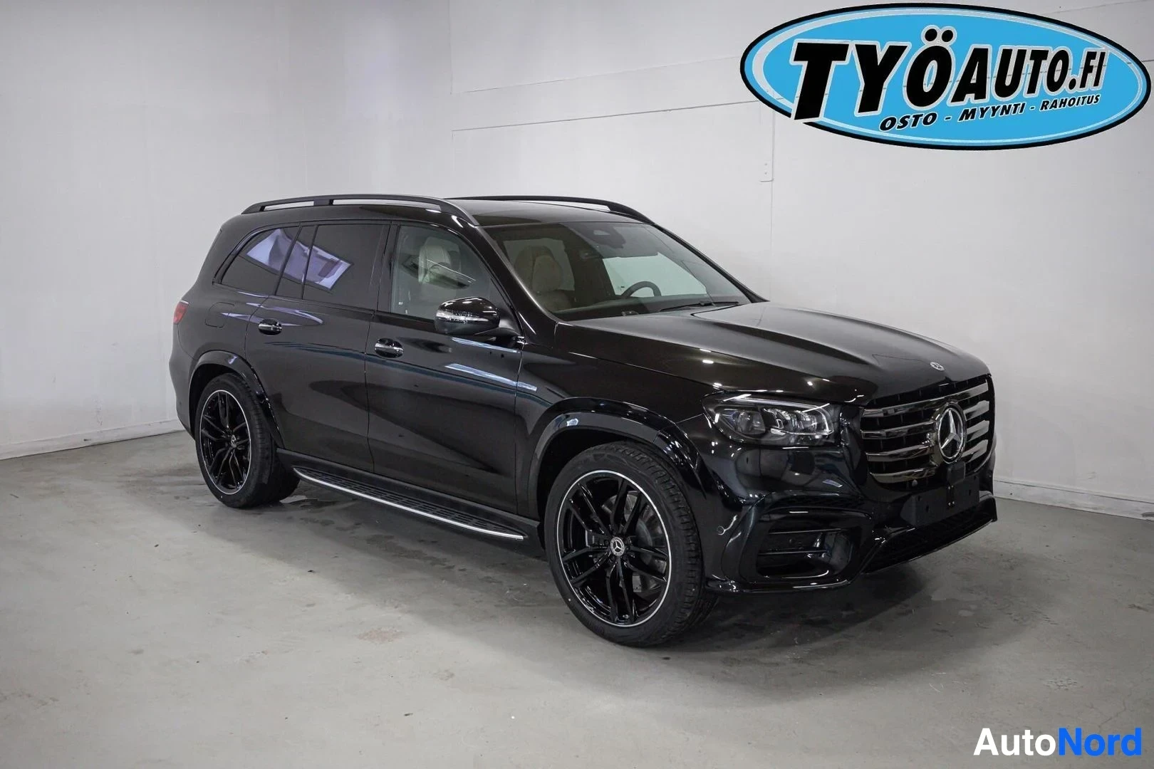 Etuviisto, nokka oikealle: Musta Mercedes-Benz GLS SUV edestä ja oikealta sivulta kuvattuna valkoisessa hallissa. Autossa on mustat monipuolaiset vanteet ja kattokaiteet.