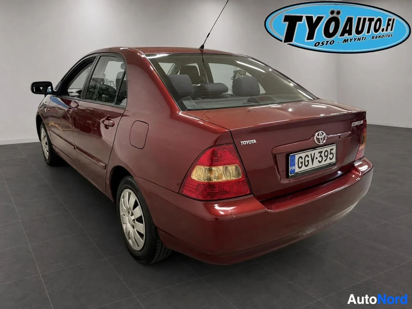 Takaviisto: Punainen henkilöauto takaviistosta kuvattuna, ajovalot ja osa kylkeä näkyvissä. Auton takaluukussa on Toyota-logo ja rekisterikilpi tekstillä. Oikeassa