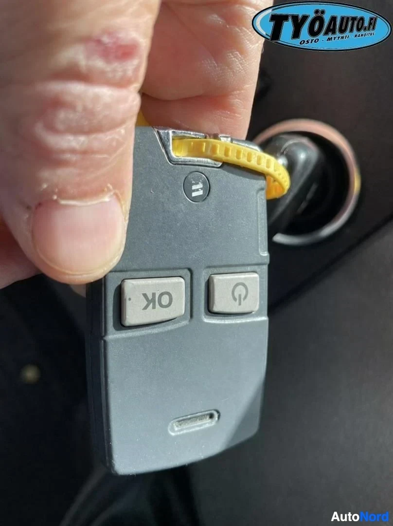 Yksityiskohta: Harmaa avainperä, jossa on kaksi nappia teksteillä "OK" ja virtasymboli. Avainperän yläosassa on keltainen rengas ja logo, jossa lukee "TYÖAUTO.fi".
