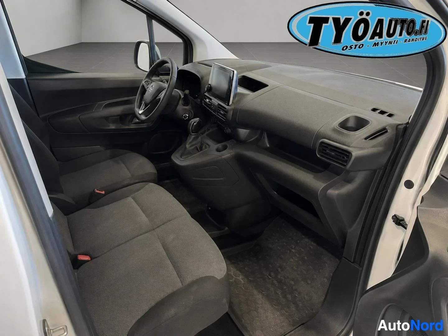 Ohjaamo: Kuvakulma auton oikean etuoven suuntaan, sisätilat näkyvissä. Kaksi kangasverhoiltua istuinta edessä, kojelauta ja ohjauspyörä näkyvissä. Keskikonsolissa on näy