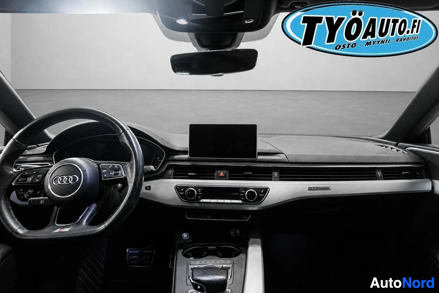 Ohjaamo: Auton ohjaamo edestäpäin. Mustassa ohjauspyörässä on Audi-logo ja monitoiminäppäimet. Keskikonsolissa on näyttö ja ilmastoinnin säätimet. Vaihdevalitsin on kesk