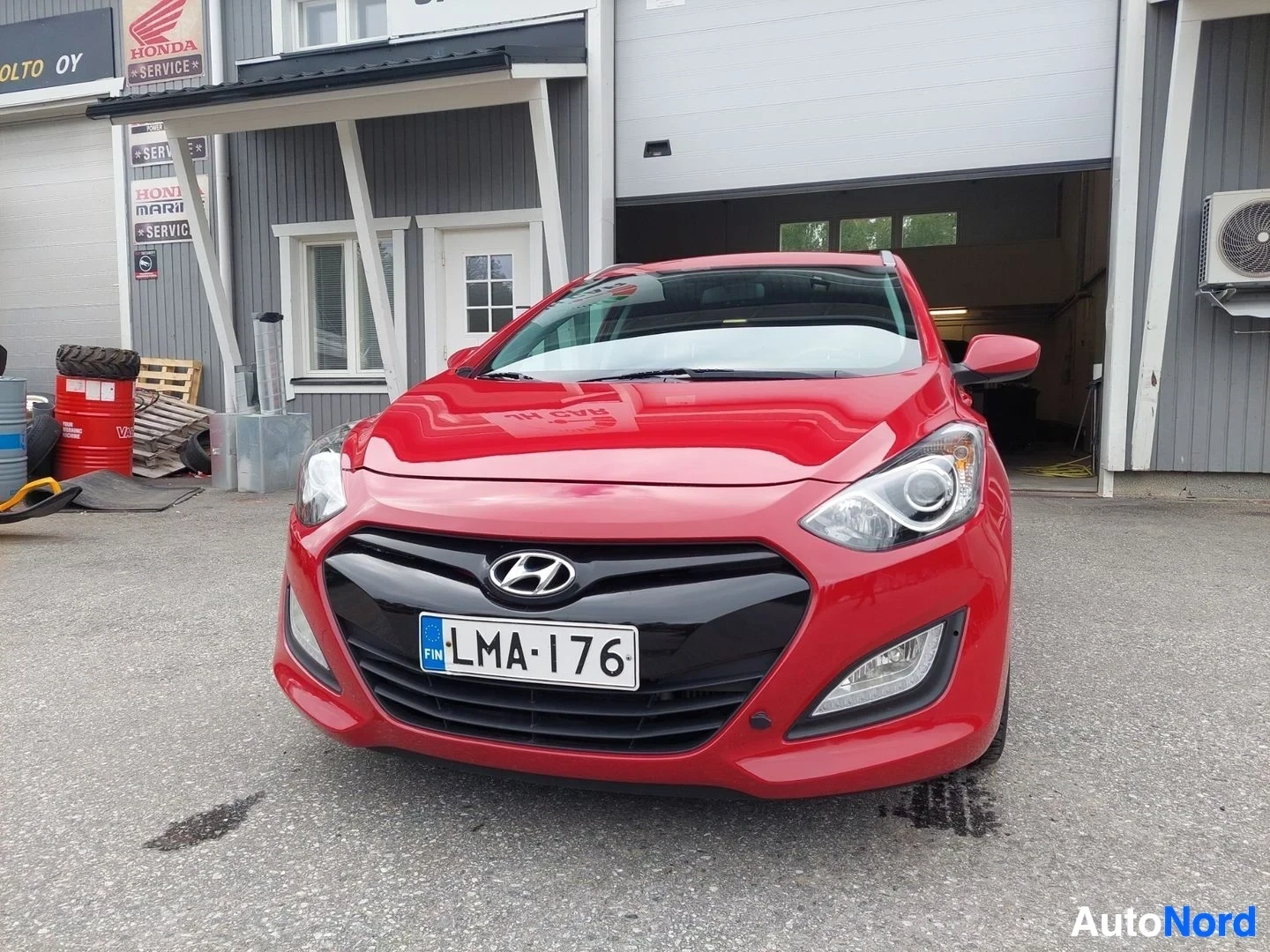 Edestä: Punaisen Hyundai i30 farmarin etuosa. Autossa on rekisterikilpi ja etusumuvalot. Rekisterikilven ympärillä on musta kehys